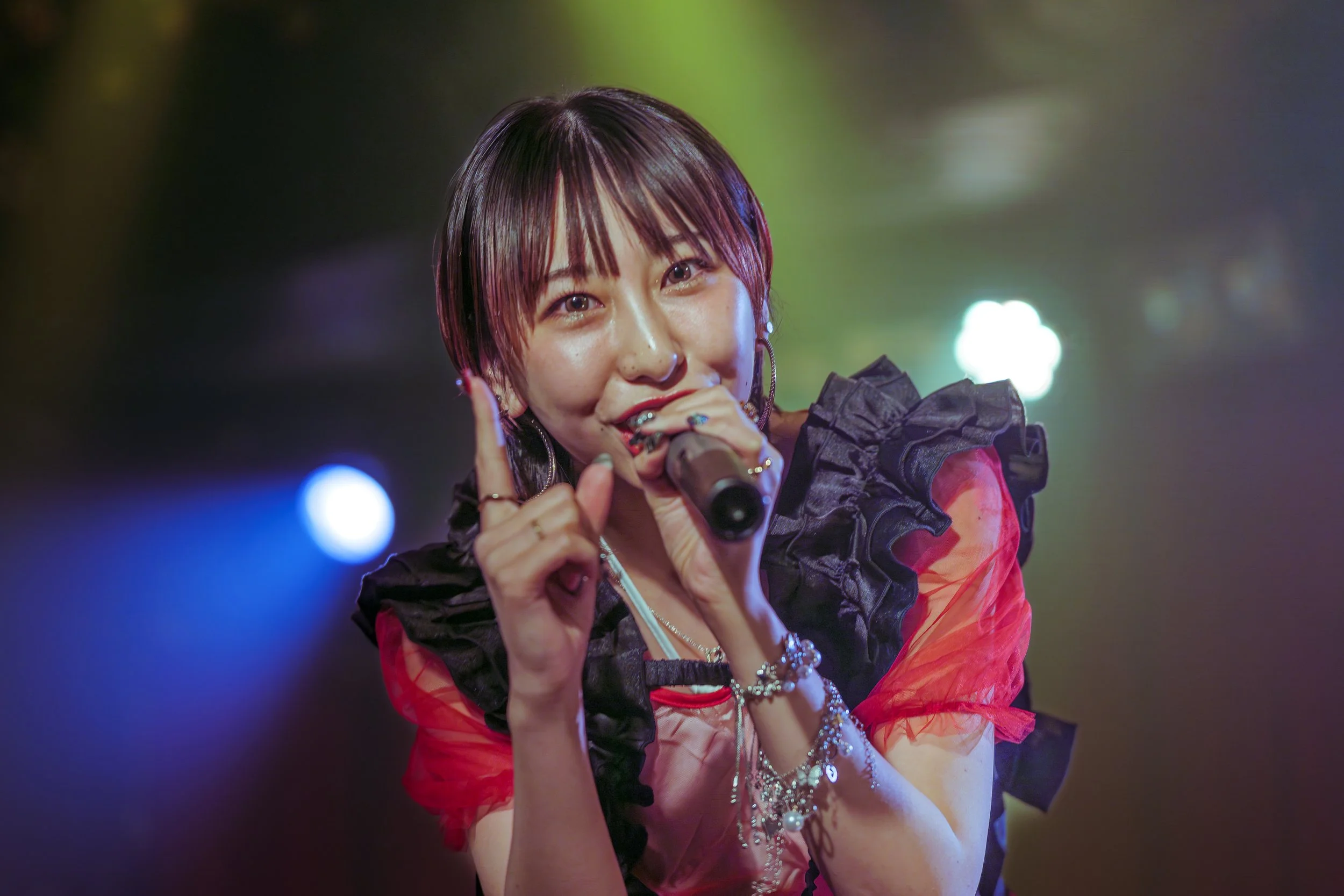 マイクを持ちながら歌う女性アイドル歌手のアップショット。黒と赤の衣装と暗い背景にキラキラしたライト。