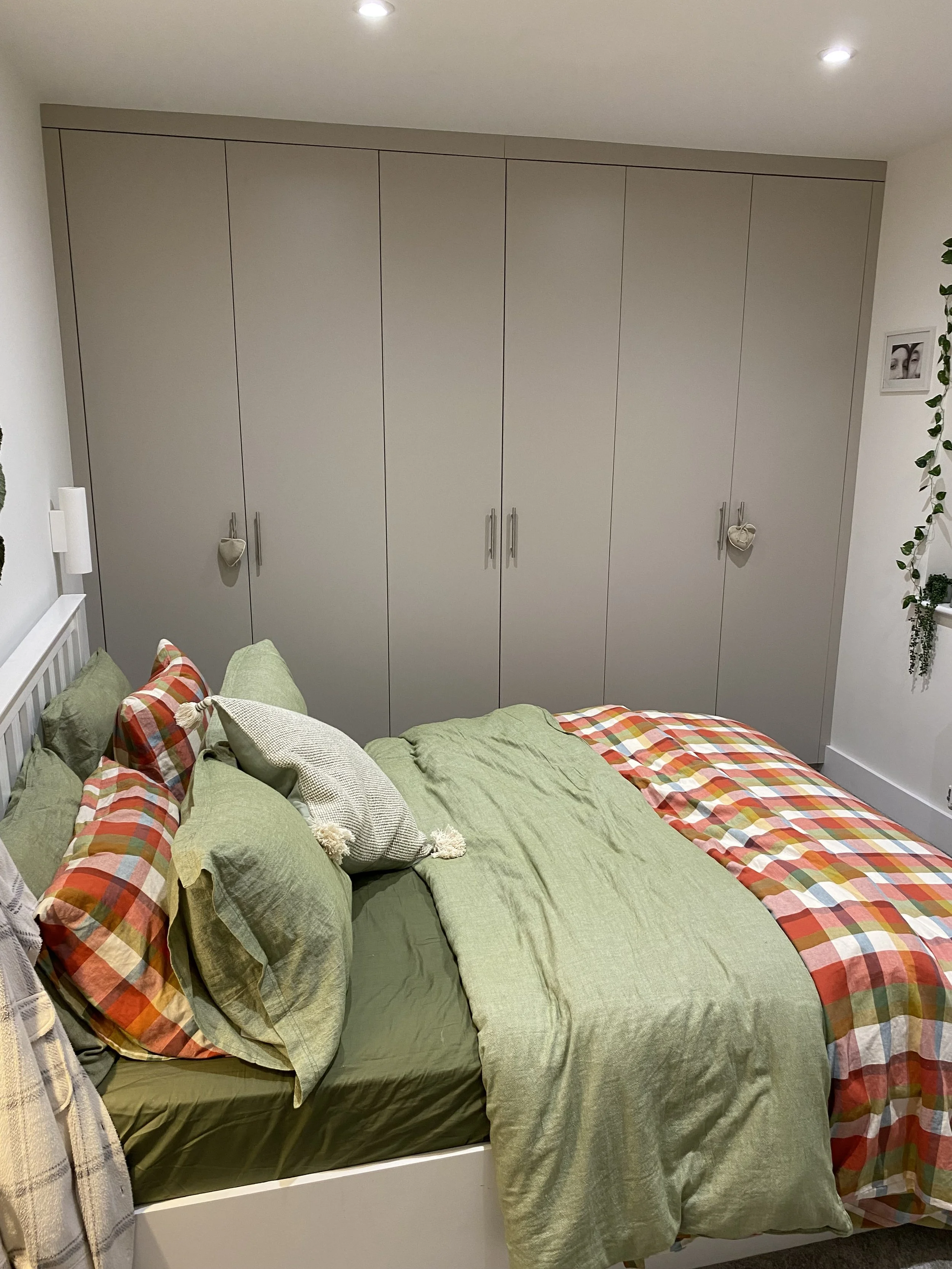 A neatly made bed with green sheets and multicolored plaid and solid pillows in front of a large beige closet with two hanging decorative hearts.