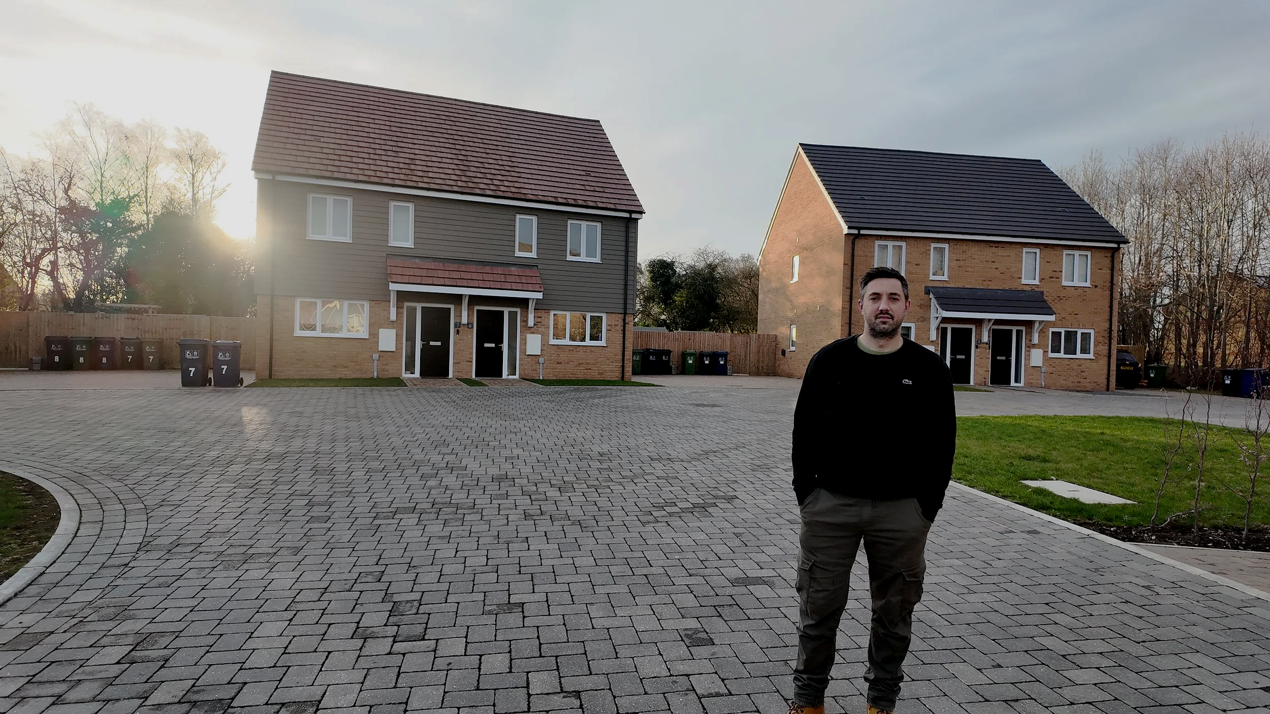 A man standing on a paved driveway in front of two new houses during sunset