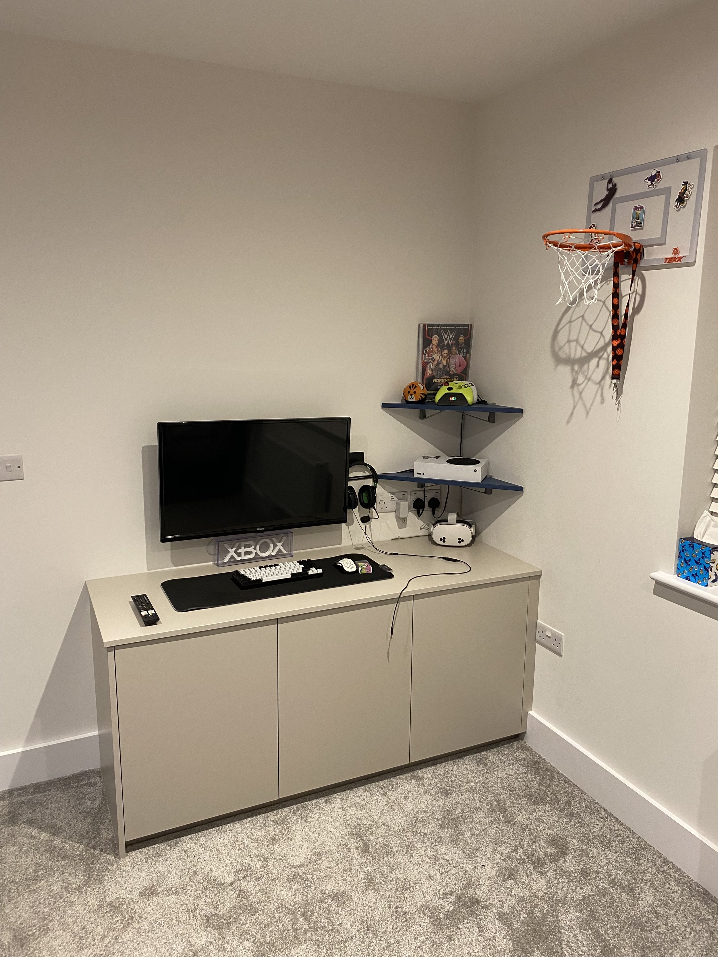 A white cabinet holding a black television, gaming accessories, and a remote, with wall-mounted shelves above that have gaming gear, a WWE poster, and a miniature basketball hoop with a hanging lanyard. The room has beige carpeting and white walls.