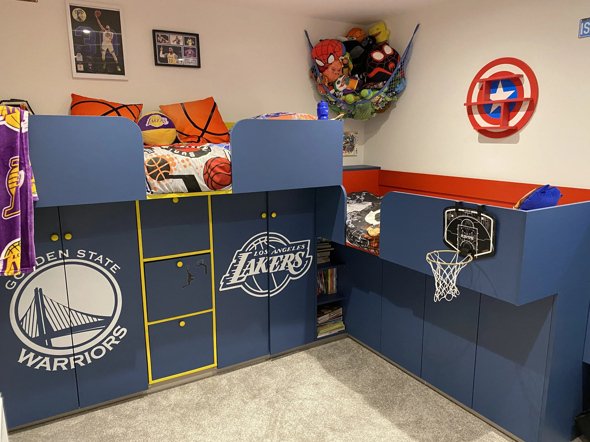 Children's bedroom with basketball-themed decor, including a loft bed with playoff pillows, sports posters, a small basketball hoop, and a wall shelf with plush toys and masks.