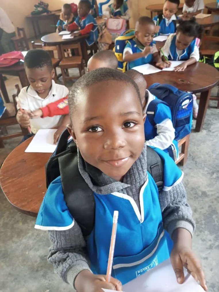Young boy smiling at camera in classroom, holding pencil and paper, with other children in background working at desks.