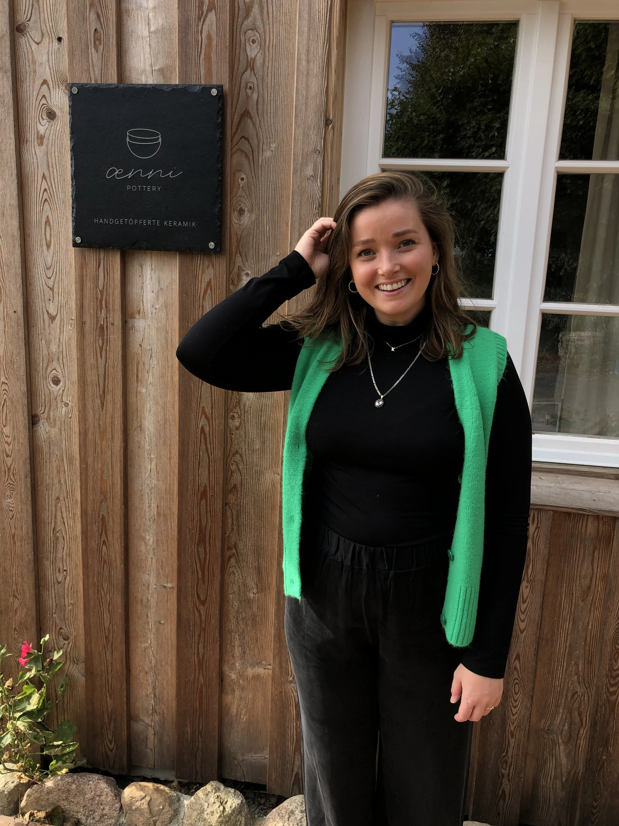 Eine lächelnde Frau steht vor einem Holzhaus, neben einem dunkelgrauen Schild, das mit weißer Schrift 'aemi pottery' und 'Handgefertigte Keramik' beschreibt.
