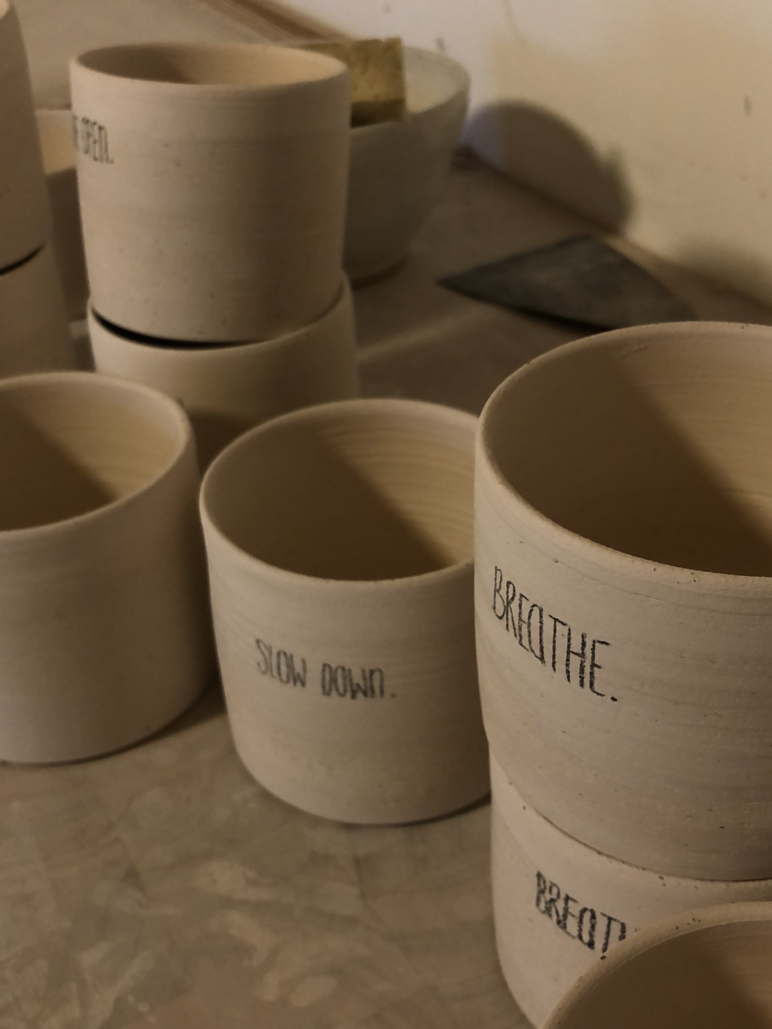 Mehrere unglasierte Keramiktassen mit handgeschriebenen Worten wie "SLOW DOWN," "BREATHE," auf einem Tisch.