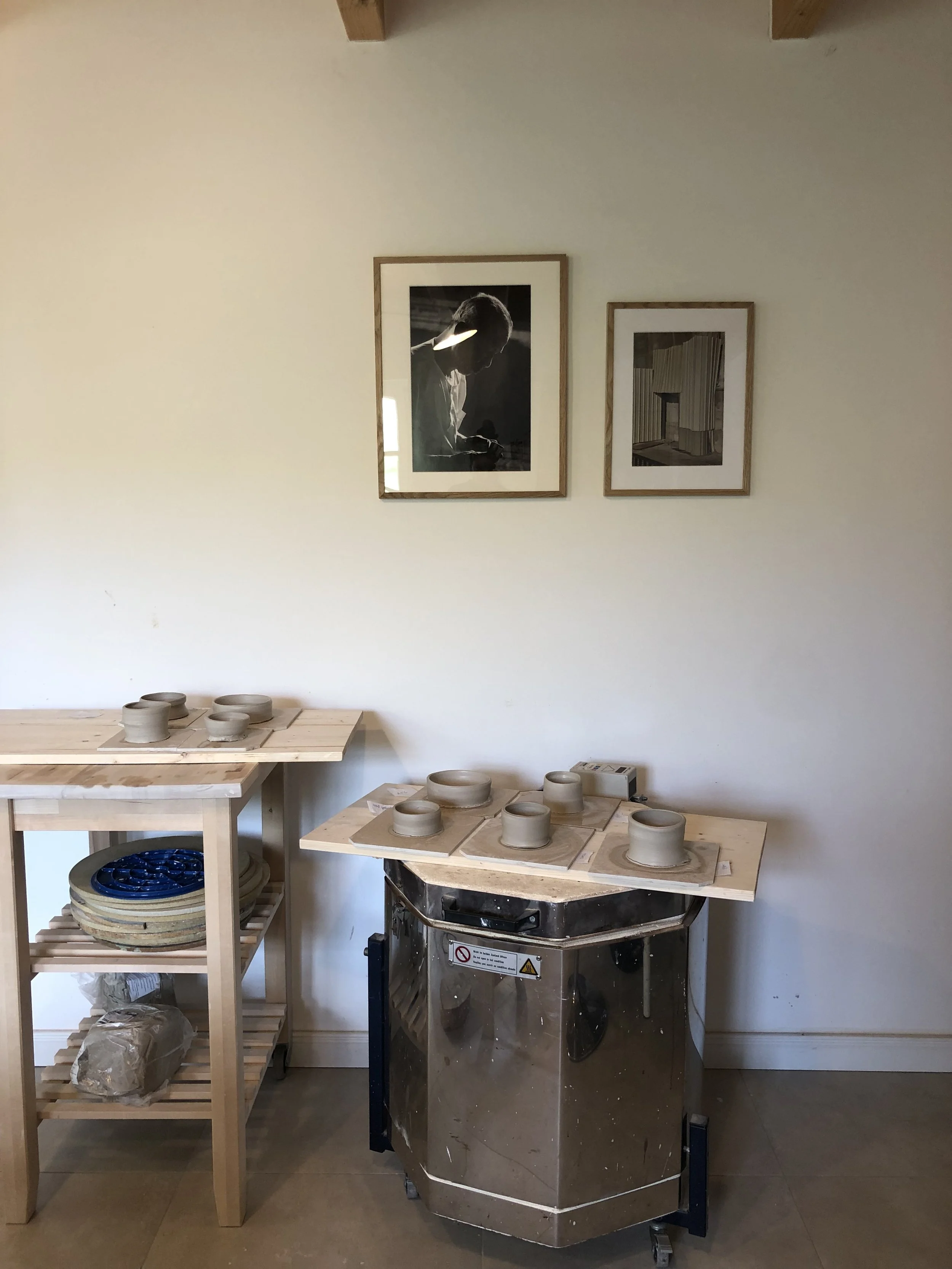 Töpferwerkstatt mit mehreren unbearbeiteten Keramikgefäßen auf Tonpergamenten und einem Holzregal mit weiteren Keramikstücken. Im Hintergrund hängen zwei Schwarz-Weiß-Fotografien an der weißen Wand.