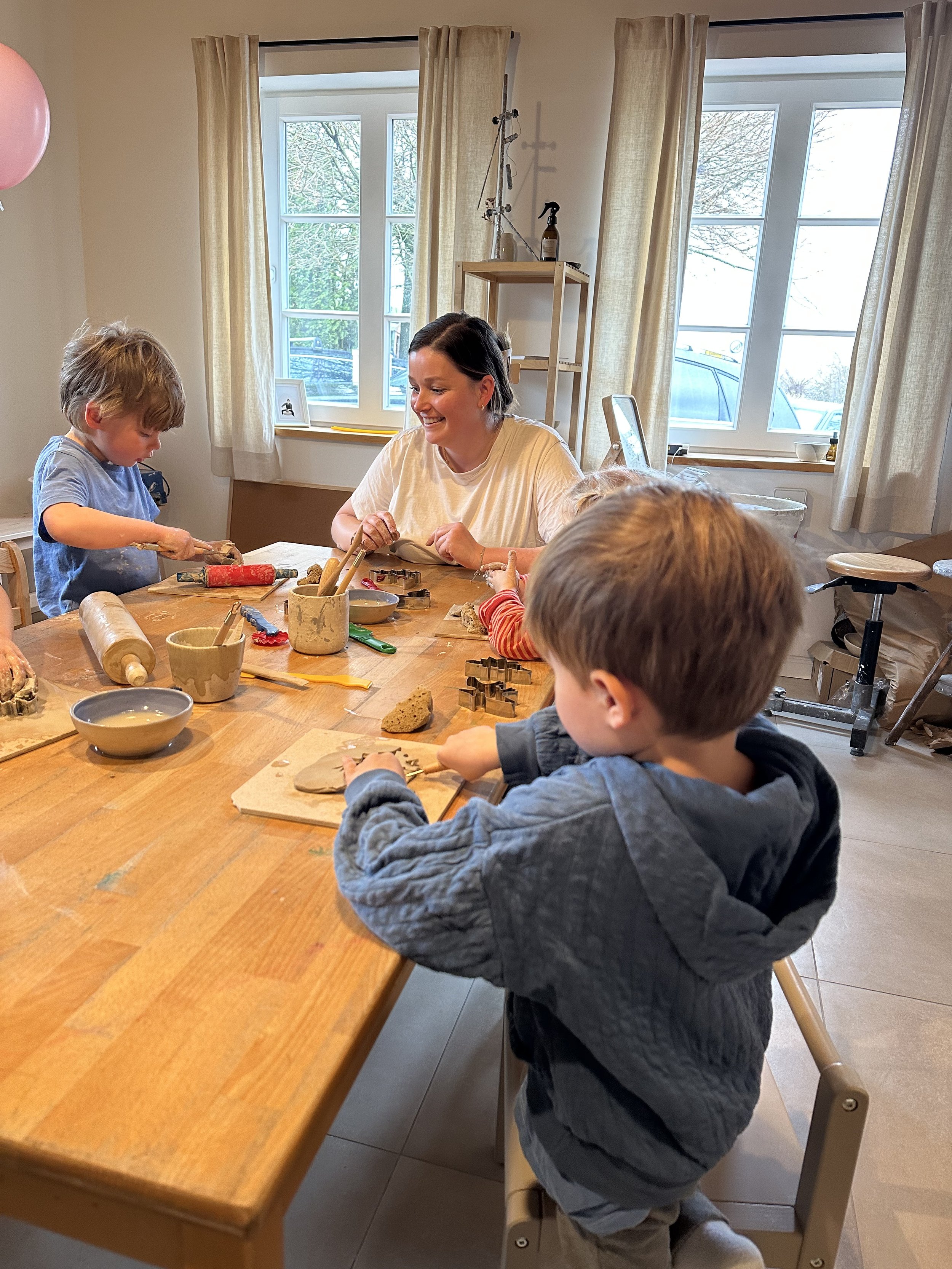 Familie backt und dekort Kekse in einer Küche mit großen Fenstern. Vier Kinder und eine Erwachsene sitzen um einen Holztisch, der mit Keksen, Teigwaren und Backutensilien bedeckt ist.