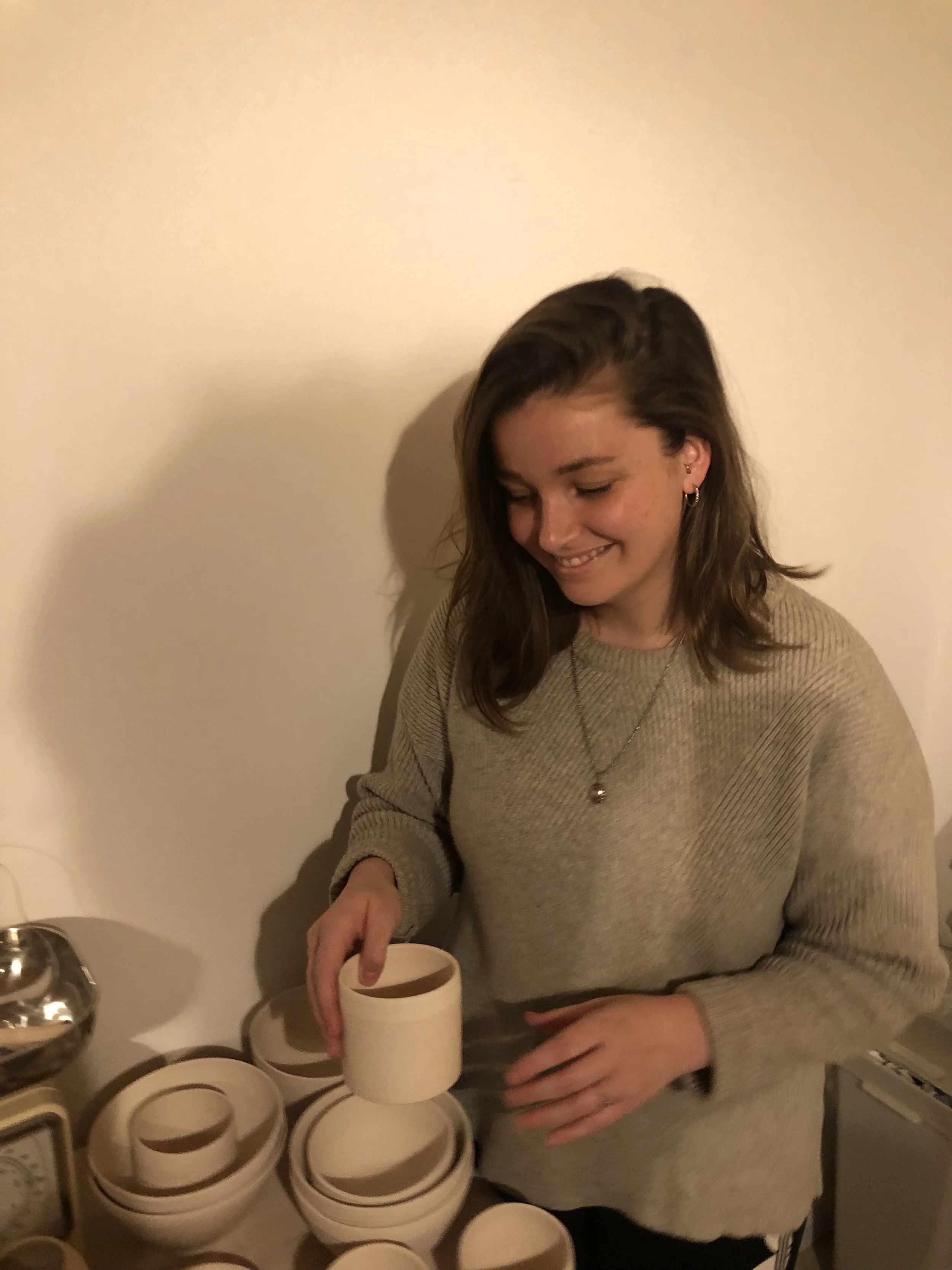 Eine lachende junge Frau mit braunen, gelockten Haaren in einem beigen Pullover, die eine weiße Kaffeetasse hält, neben mehreren weiteren Tassen auf einem Tisch.