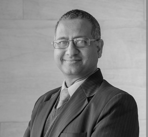 A middle-aged man in a dark pinstripe suit, blue shirt, and blue tie, smiling with short dark hair, glasses, standing indoors against a wooden wall background.