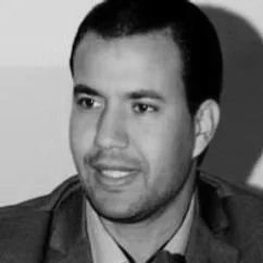 A man with short dark hair and a light brown complexion, wearing a dark blazer and light shirt, looking to the side indoors.