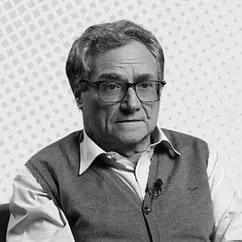 A middle-aged man with glasses and gray hair, wearing a light blue shirt and a gray sweater, looking thoughtful or serious.