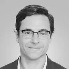 Portrait of a man wearing glasses with short, dark hair, dressed in a dark blazer and light shirt, against a light grey background.
