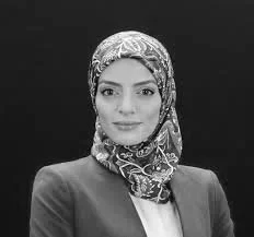 Woman wearing a colorful patterned hijab and a blue blazer against a black background.
