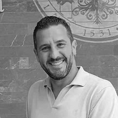A smiling man with short dark hair and a beard, wearing a light blue polo shirt, standing outdoors in front of a stone wall with a decorative clock face.