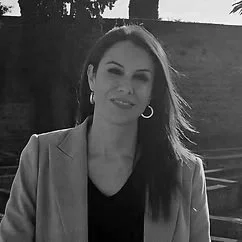 A woman with long brown hair, wearing a beige blazer and black top, smiling outdoors with trees in the background.