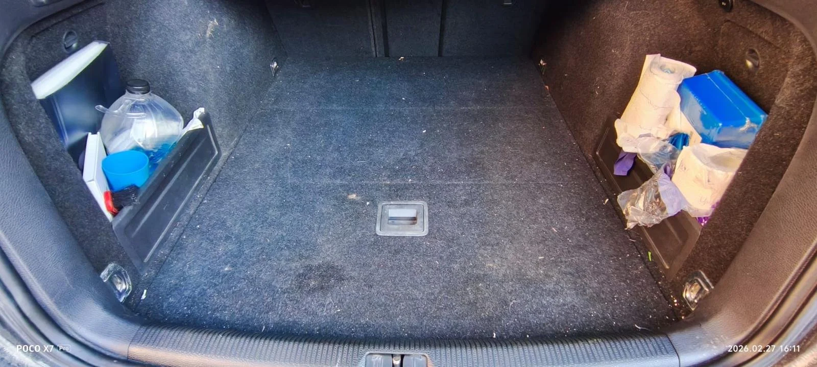 The trunk of a car contains a blue plastic water bottle, a blue plastic cup, a black item, and various packages of tissues or paper towels on the right side.