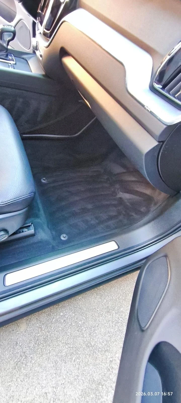 Interior of a vehicle showing the passenger side, including part of the dashboard, footwell with a rubber mat, seat, and door speaker, with an open door revealing the ground outside.