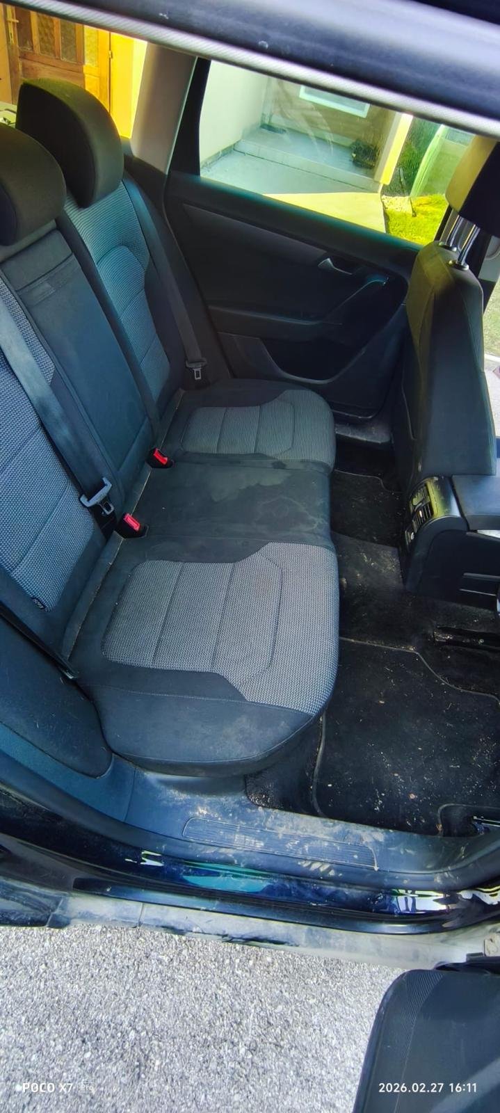 The backseat of a car showing a gray and black fabric bench seat with three seat belts, a black door panel, and a window showing a porch and a sidewalk outside.