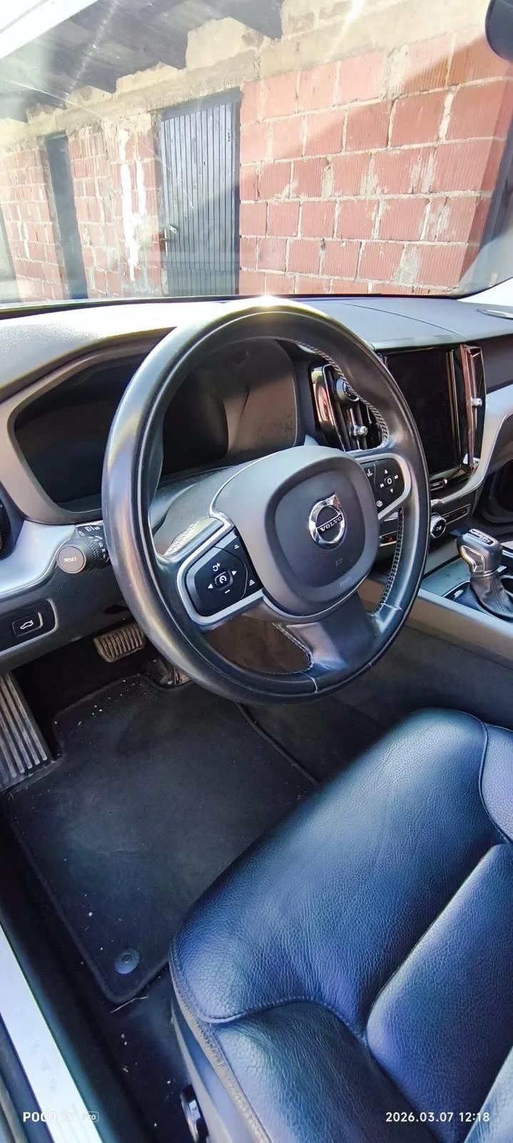 Inside view of a Volvo car showing the steering wheel, dashboard, and front seat with a brick wall and door visible through the windshield.