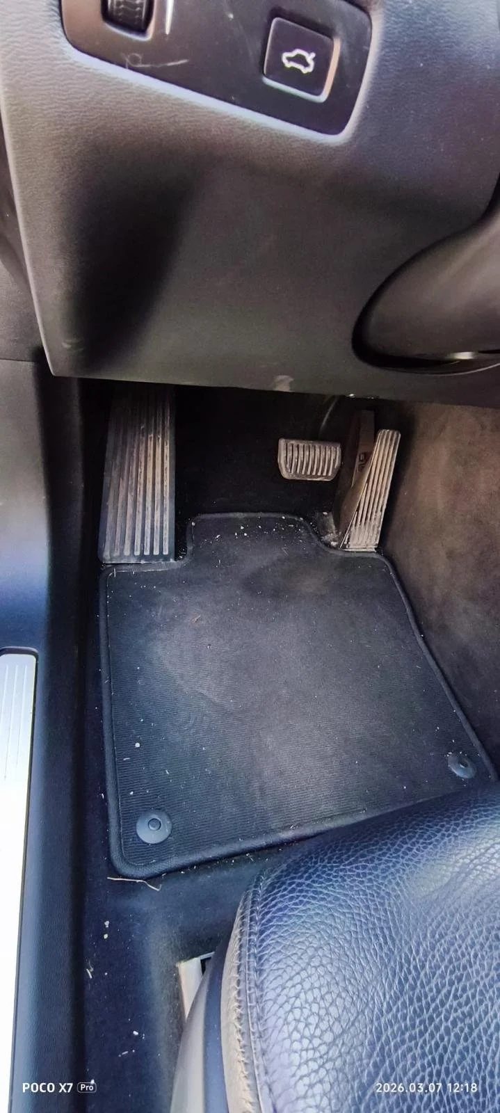 The image shows the footwell area of a car's driver's side, with the gas pedal, brake pedal, and an accelerator pedal visible. There is a black floor mat with some dirt and debris underneath the pedals and a portion of the driver's seat with black leather upholstery.