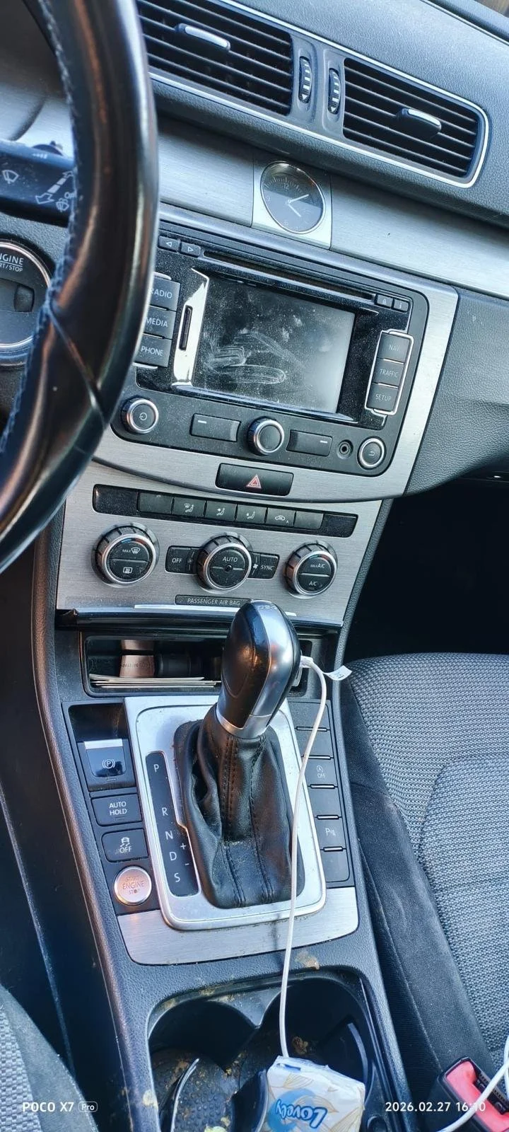Inside a car showing the dashboard, steering wheel, gear shift, and center console with various controls, a clock, and a phone charger with a connected device.