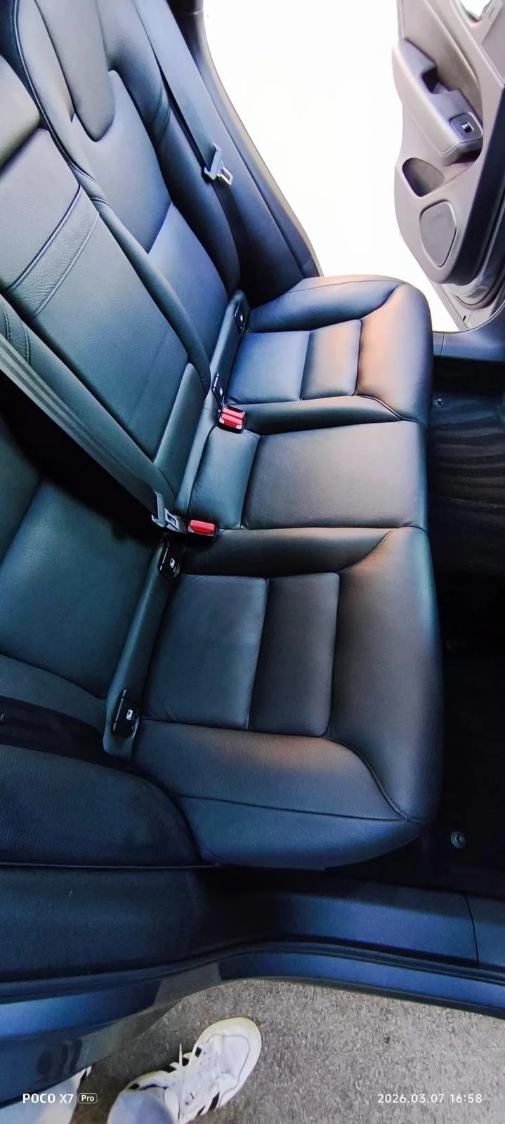 Back seat of a car with black leather upholstery, showing three seatbelt buckles and part of the open front passenger door.