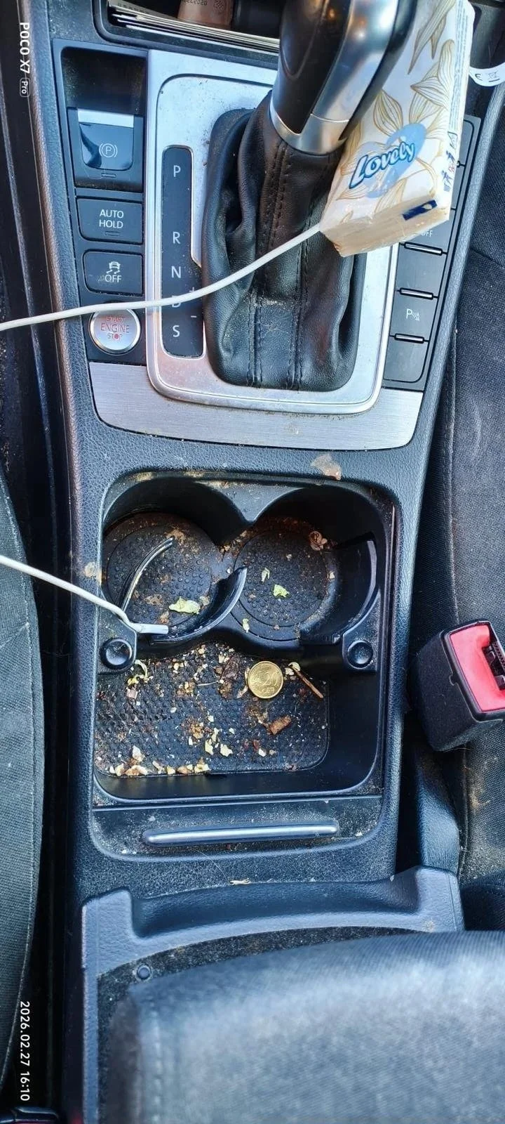 Car interior with dirty cup holder containing crumbs, a coin, and trash, next to gear shifter and control buttons.