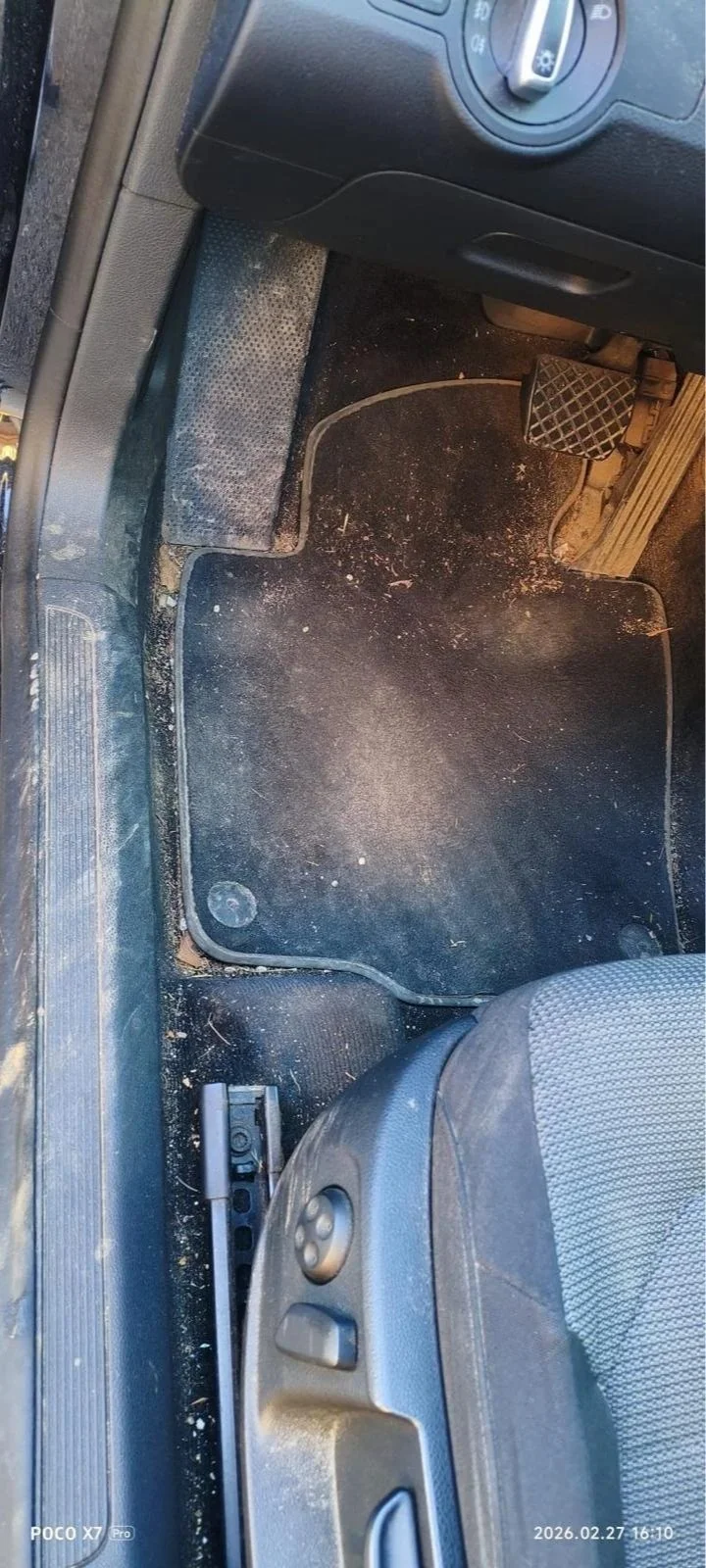 The image shows the driver's side floor area of a car, including the foot pedals, floor mat, and parts of the seat. The floor area is dusty and slightly dirty.