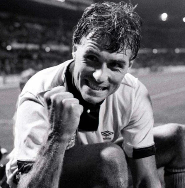 A male soccer referee celebrating with a clenched fist and a wide smile on his face, wearing a referee uniform on a soccer field.