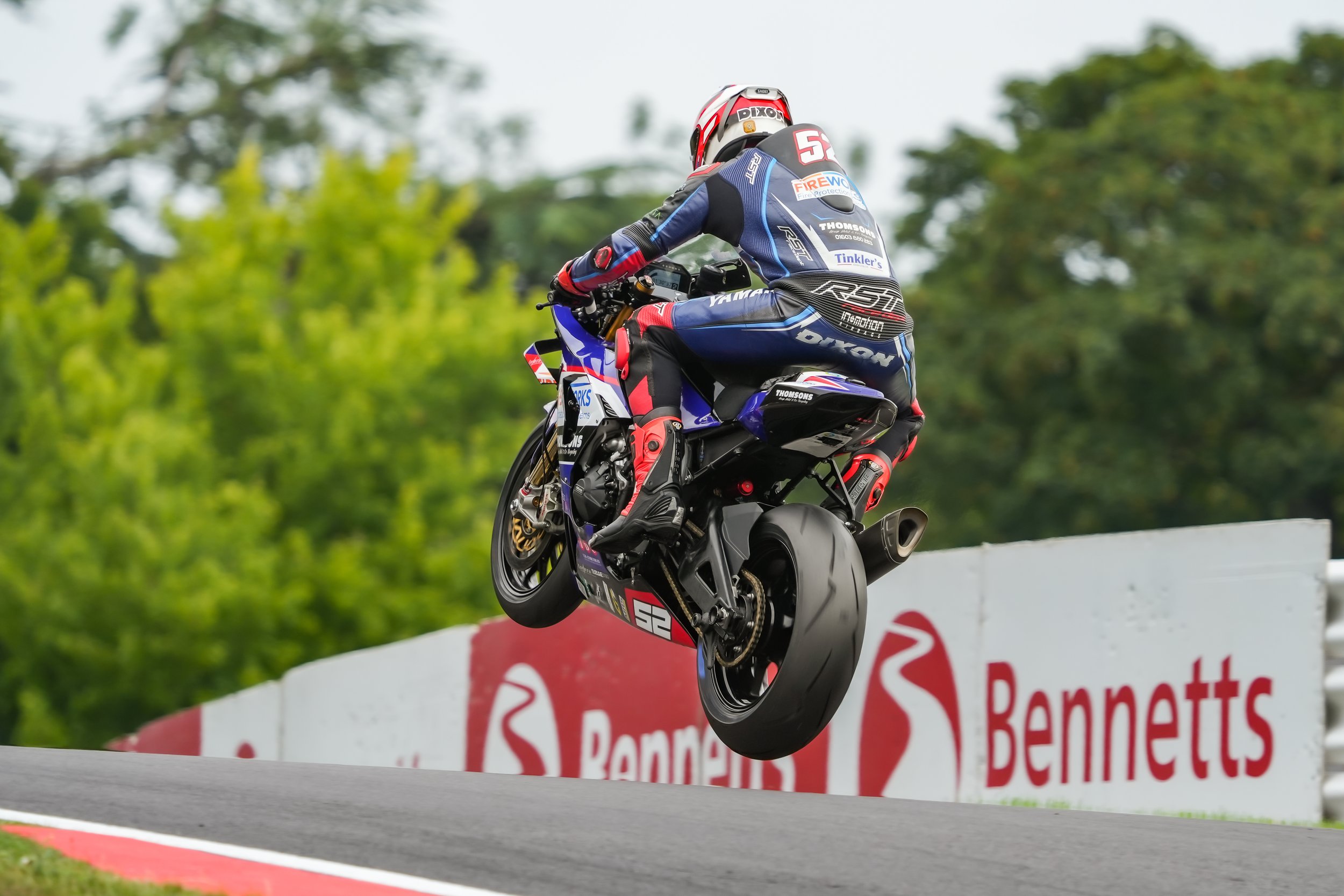A motorcycle racer jumps in the air during a race, wearing a blue and black racing suit with sponsor logos, and a helmet with red and white accents. The motorcycle is blue with the number 52, and the background features a track advertising banner wit