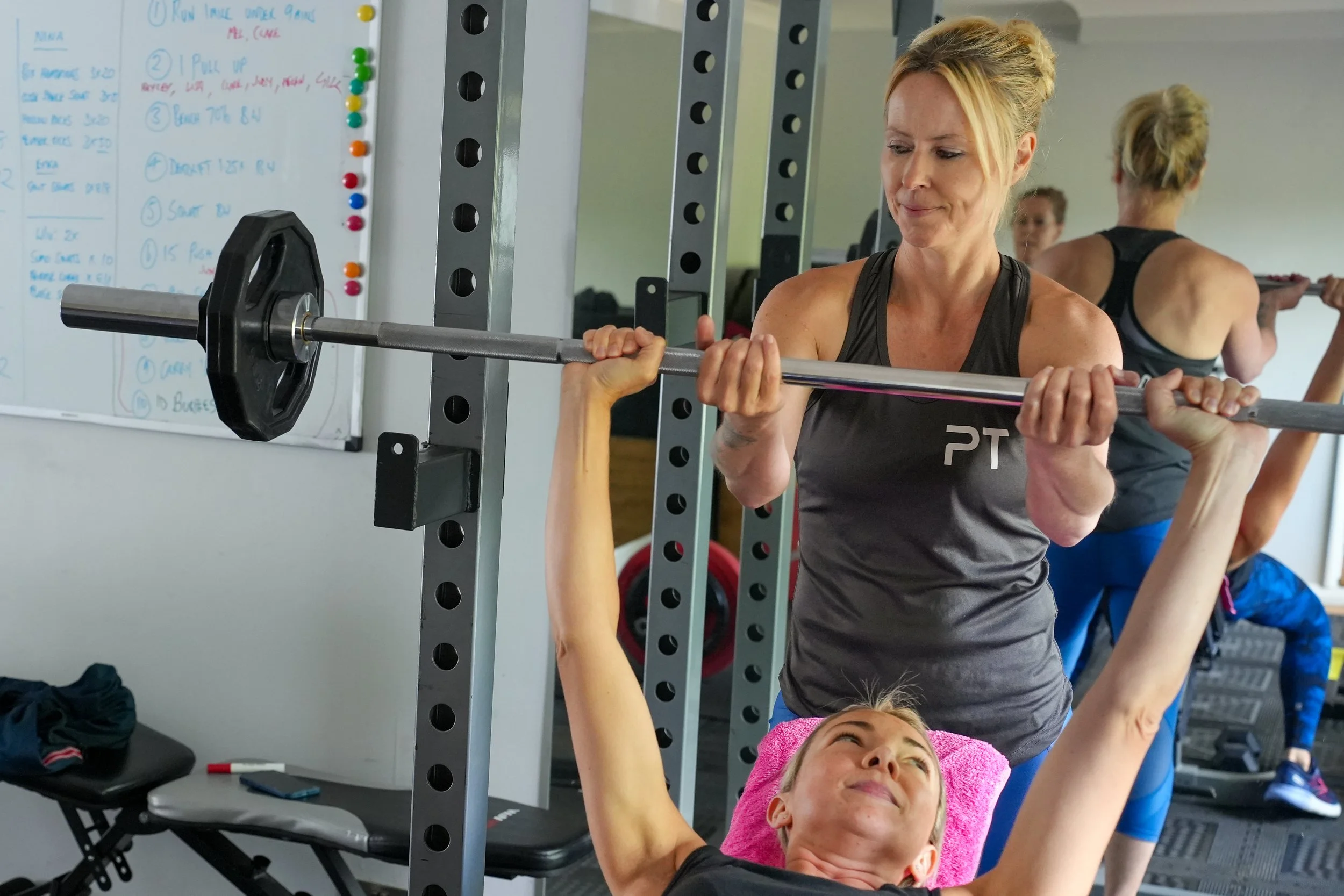 Two women in a gym, one spotting the other during a bench press exercise with a barbell. The woman lying down has a pink towel under her head and is pushing the barbell upward. The woman standing is assisting and observing. Mirrors and a whiteboard with notes are visible in the background.