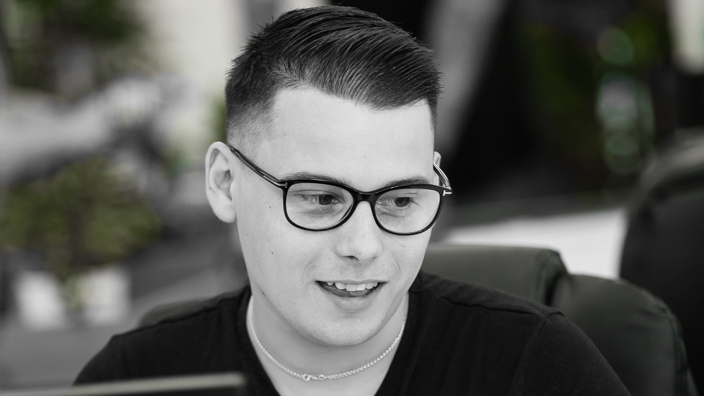 A young man with short dark hair, wearing glasses, a black t-shirt, and a silver necklace, is looking at a computer screen.