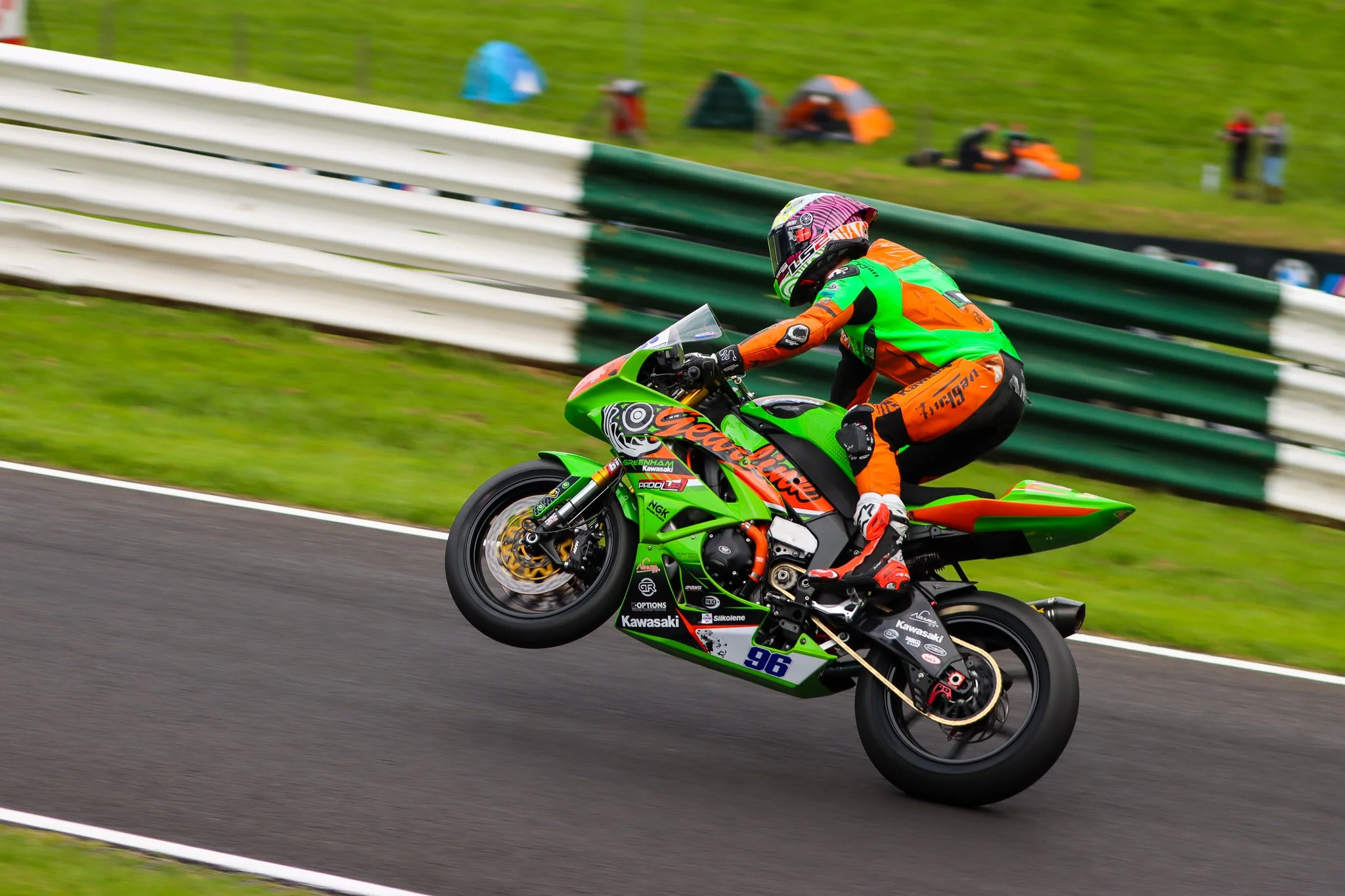 A motorcycle rider in bright orange and green racing gear performs a wheelie on a Kawasaki racing motorcycle on a race track, with tents and spectators in the background.
