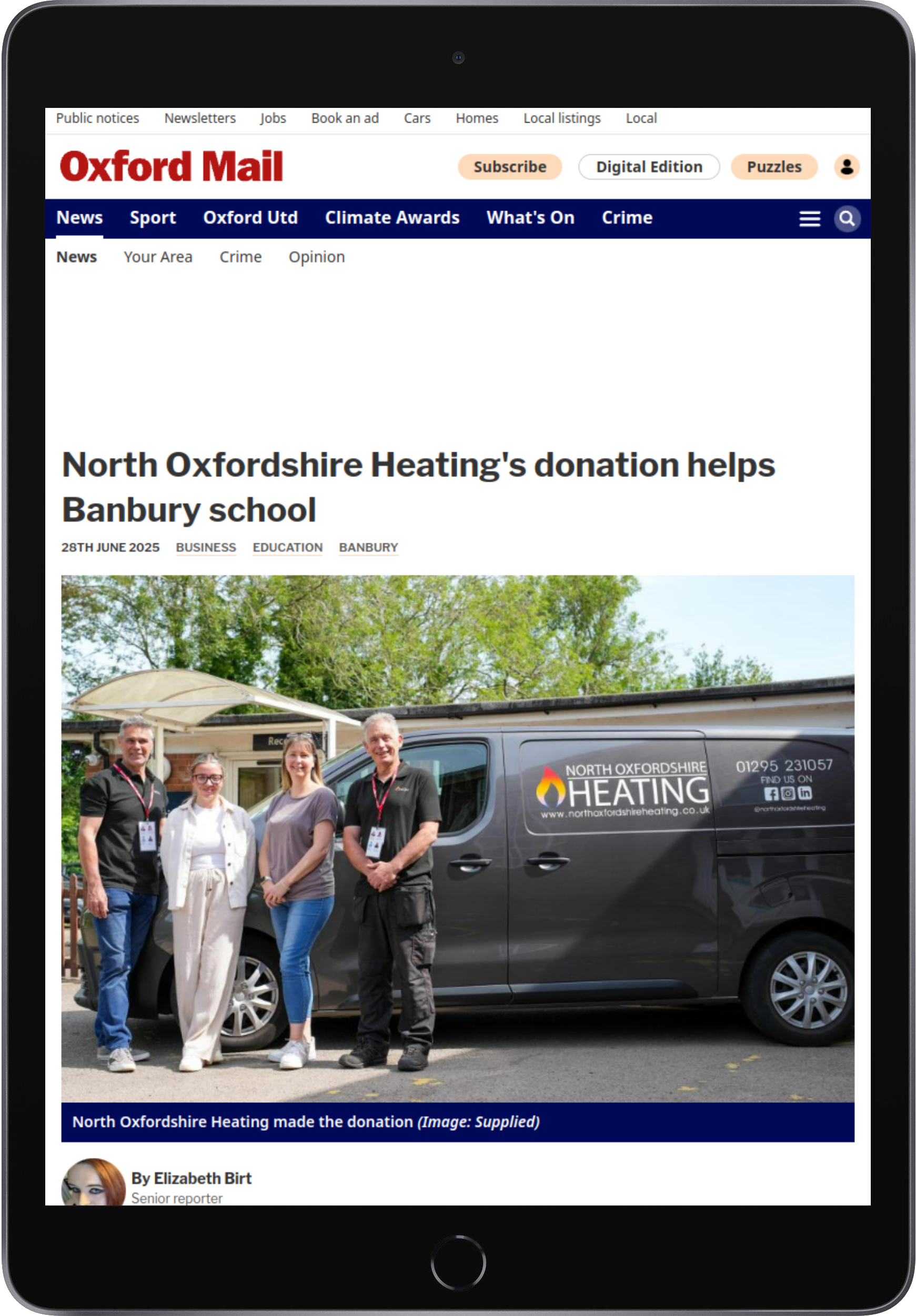 A news article on a tablet screen showing a photo of four people standing in front of a North Oxfordshire Heating van, which has the company's logo and contact information, during a donation event at Banbury School.