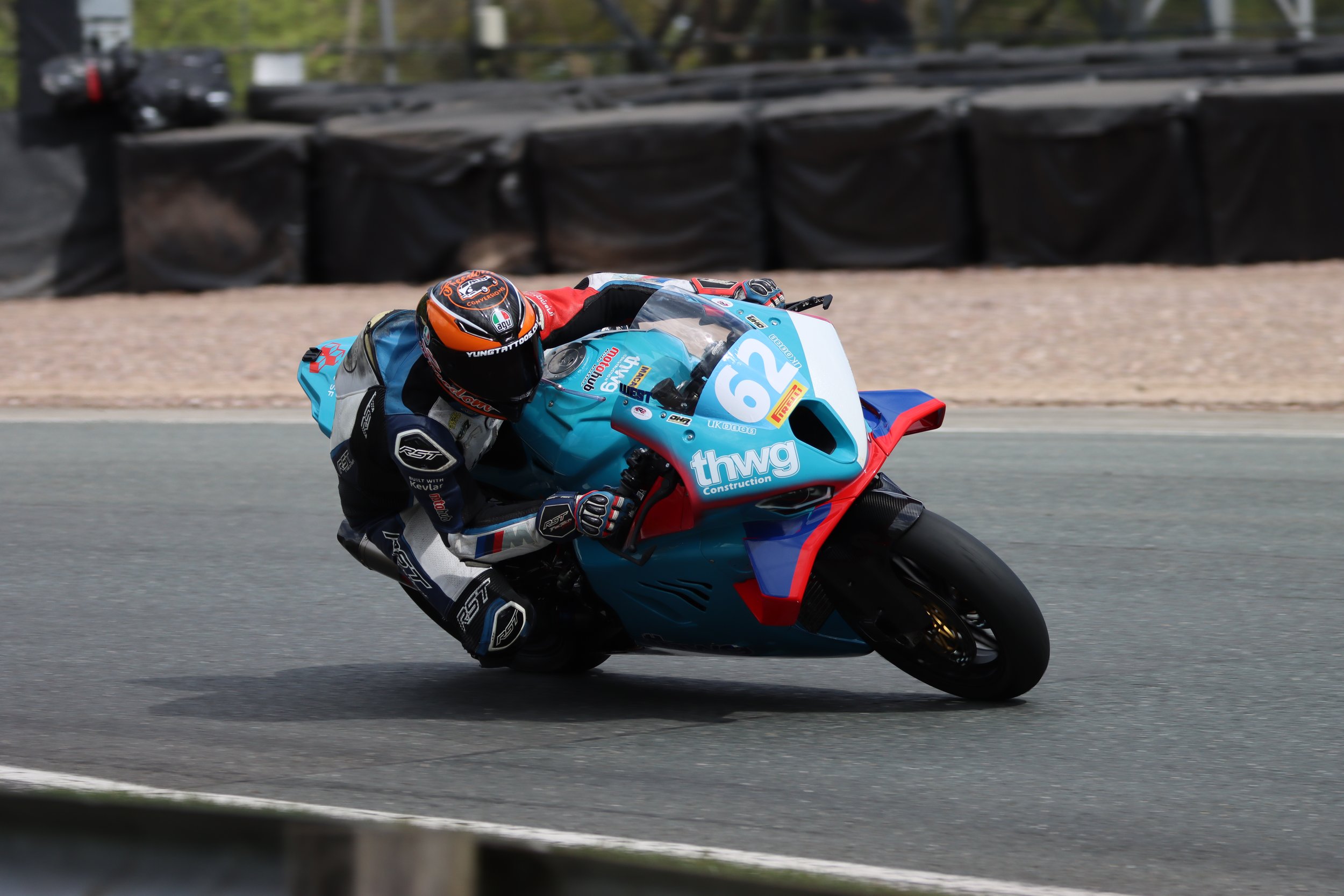 Motorcycle racer in full gear leaning into a turn on the racetrack, riding a blue and red motorcycle with number 62 and sponsor logos.