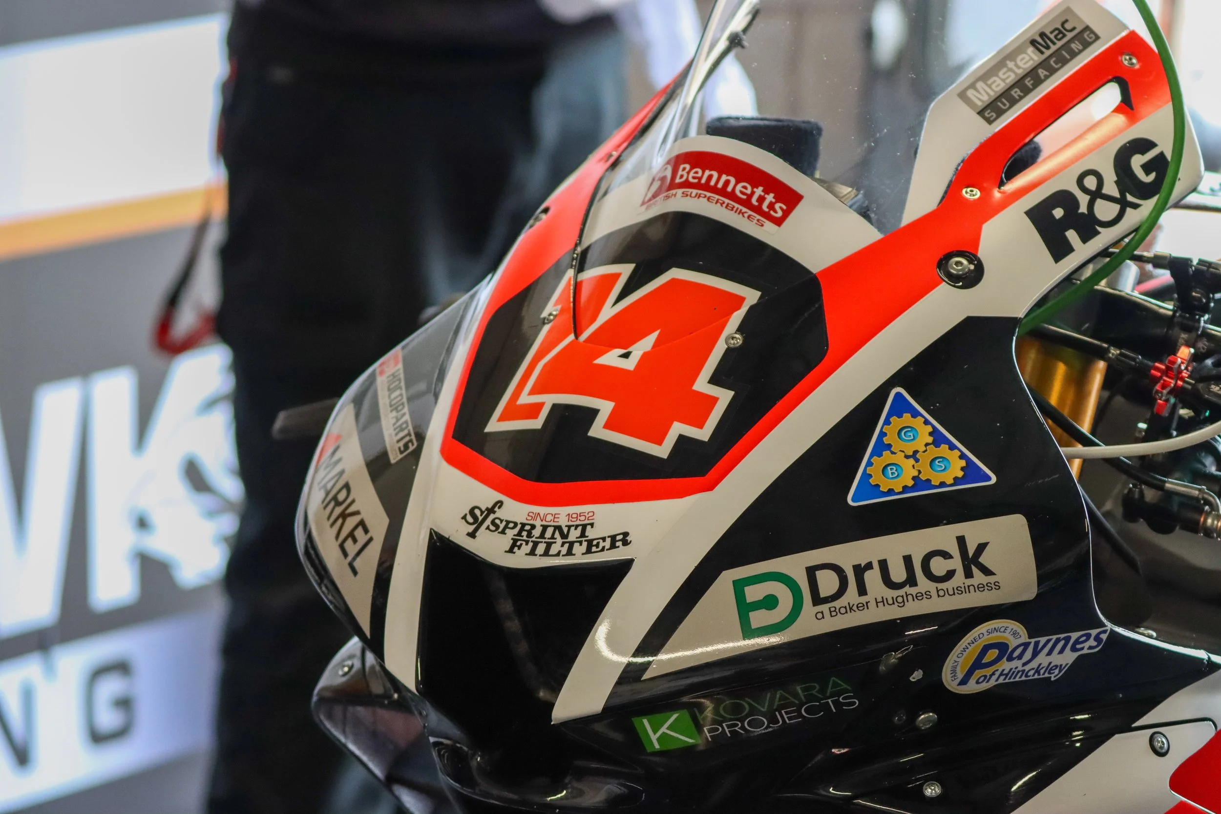 Close-up of a racing motorcycle with various sponsor logos, including a large number 4 on the front, black, white, red, and green colors, and labeled with brands like R&G, Druck, Paynes of Hinckley, and others.