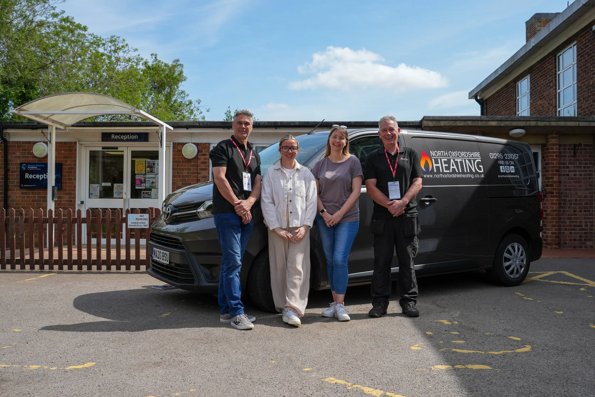 Four people standing in front of a black heating services van outside a brick building with a reception sign. The group includes two men and two women, all smiling, wearing casual attire. The van has the logo and contact information of North Oxfordsh