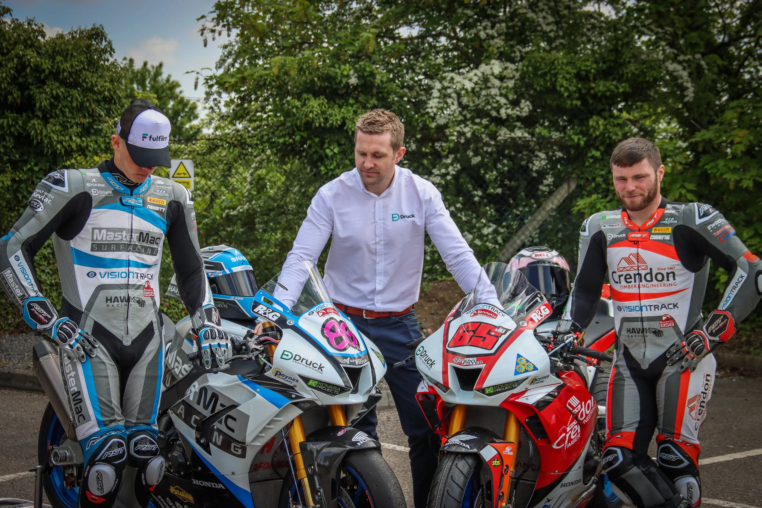 Two motorcycle racers in racing suits standing next to racing motorcycles, with a man in a white shirt inspecting the bikes outdoors