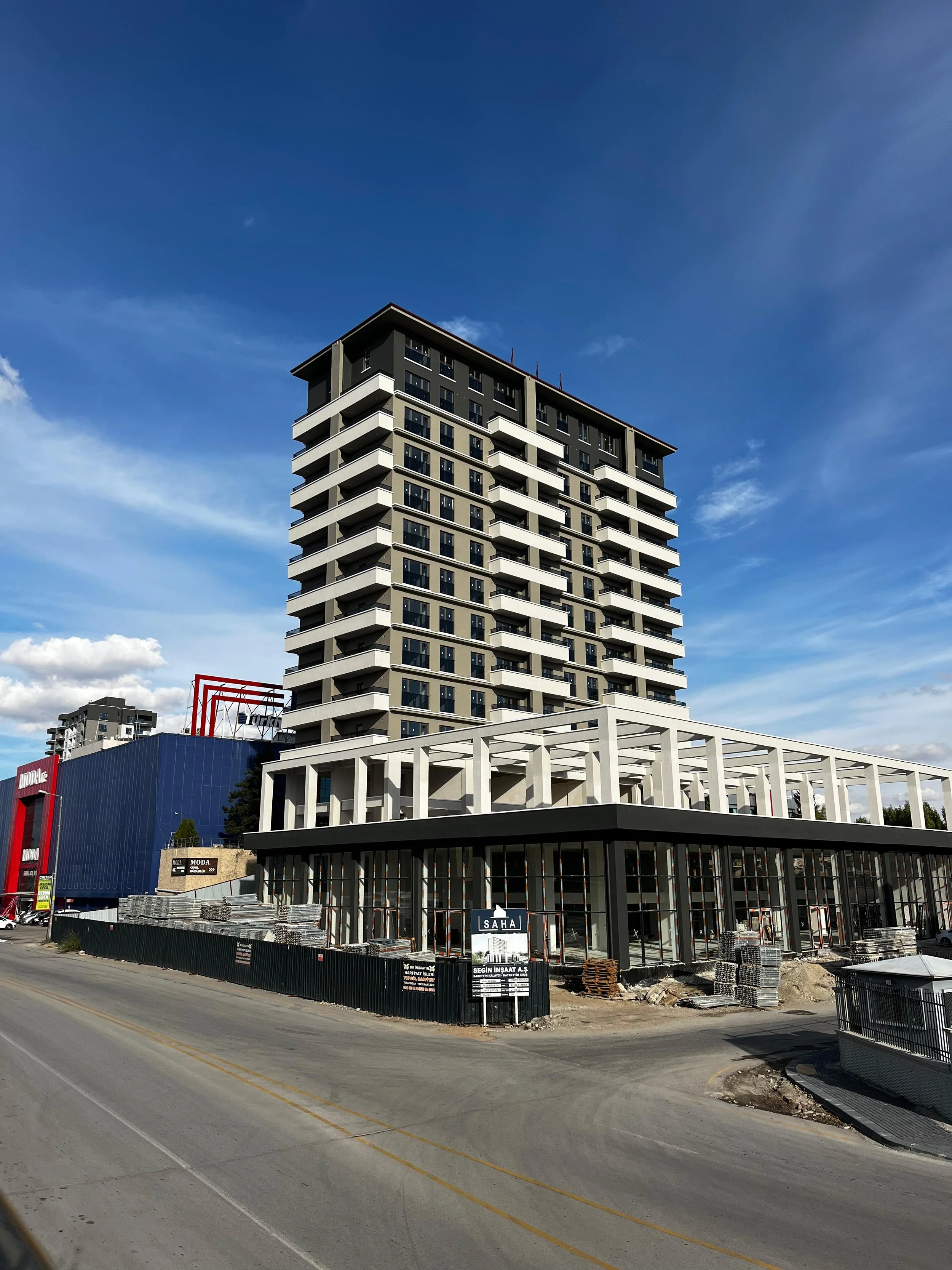 A tall, modern multi-story building under construction with a mixed-use design, featuring a retail or commercial space at the ground level and residential or office units above. The building has white and black accents, with glass windows and a construction site with materials and fences in the foreground.