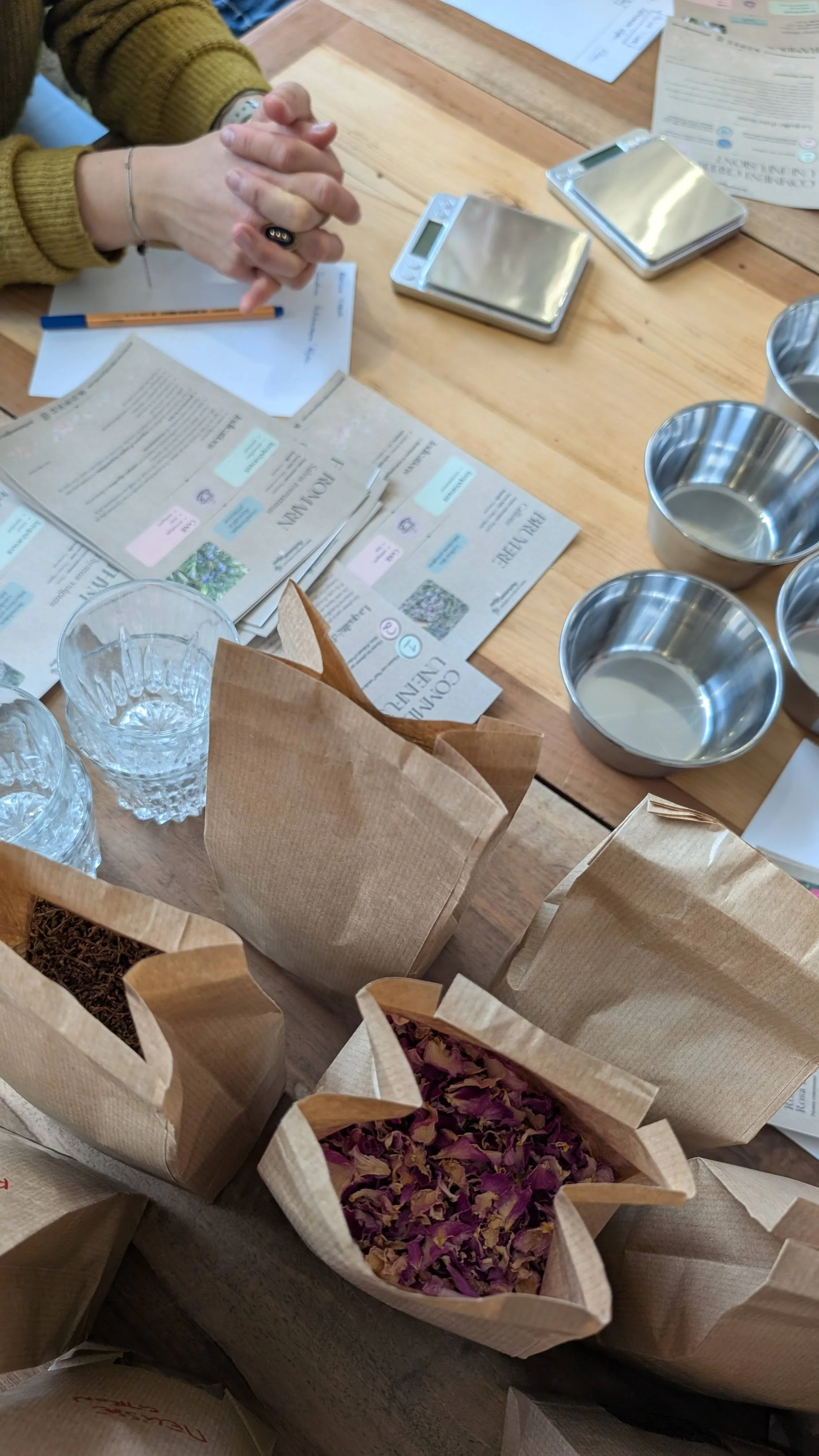 Table avec sacs en papier contenant des fleurs séchées, verres en cristal, comprimés de papier et balances électroniques.