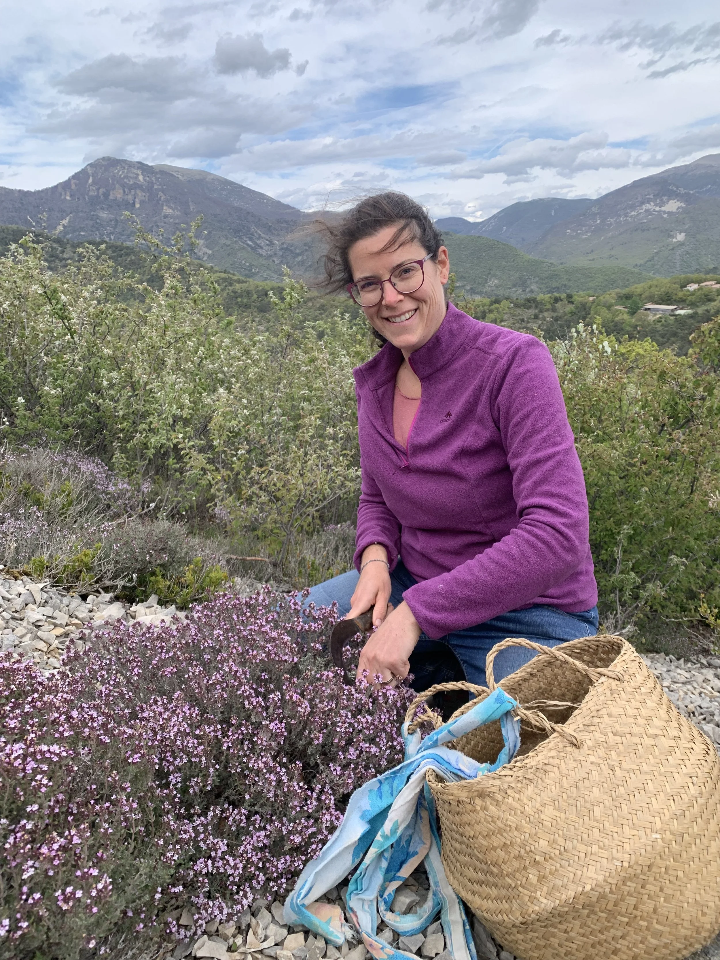 Une femme en extérieur, recueillant des fleurs de couleur rose dans un paysage de montagne avec des buissons verts, sous un ciel nuageux.