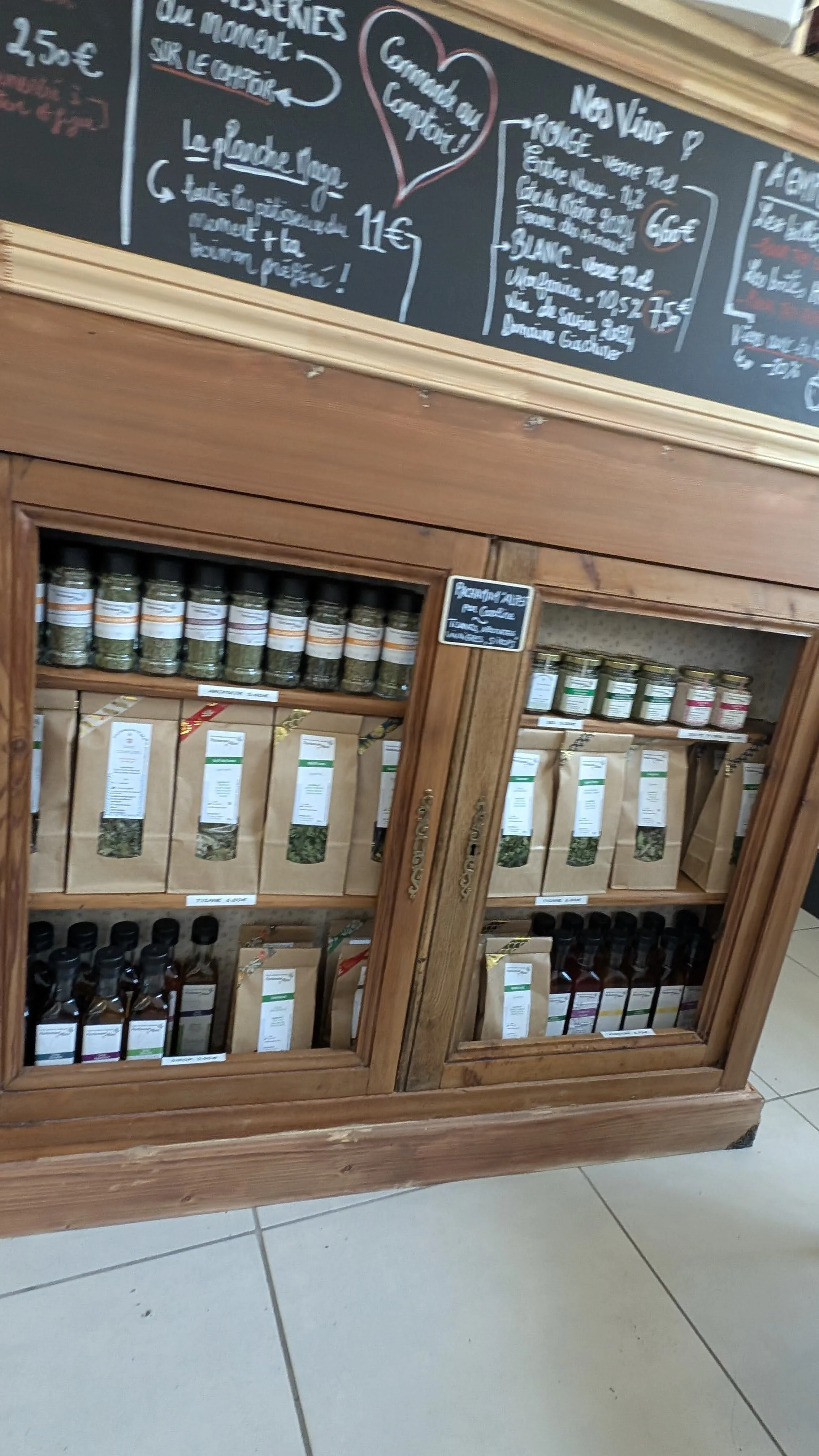 Rayonnage en bois avec des bocaux et sachets d'herbes aromatiques pour cuisine, devant un tableau noir avec menu écrit à la craie.