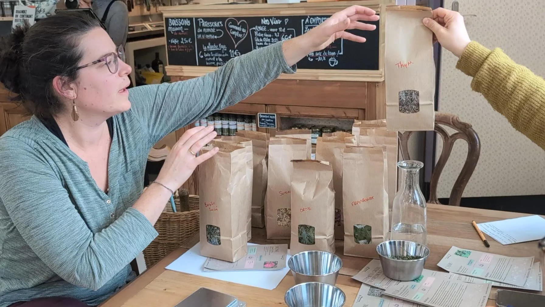 Une femme échangeant un sac en papier avec une autre personne lors d'une dégustation ou d'une vente de produits artisanaux. Plusieurs sacs en papier sont sur la table, contenant probablement des herbes ou des épices, avec des étiquettes écrites à la 