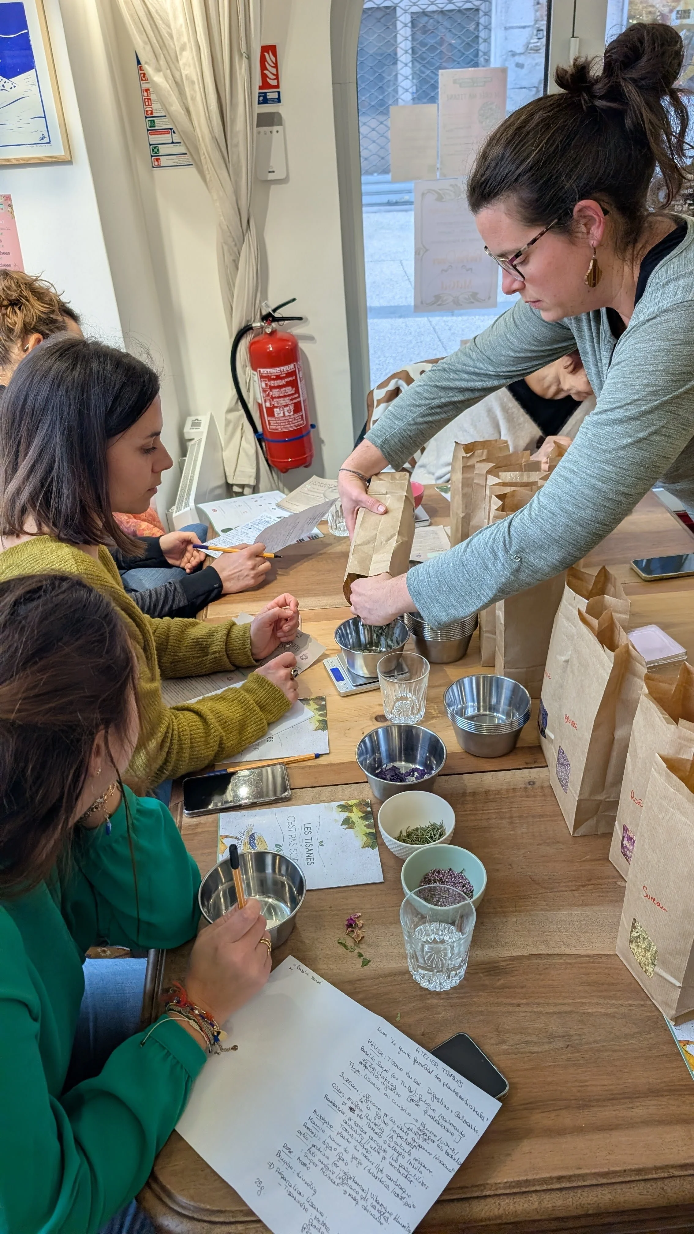 Groupe de personnes participant à un atelier de fabrication de tisane, avec des sachets en papier, des contenants métalliques, des feuilles de plantes séchées, et des documents écrits sur la table.