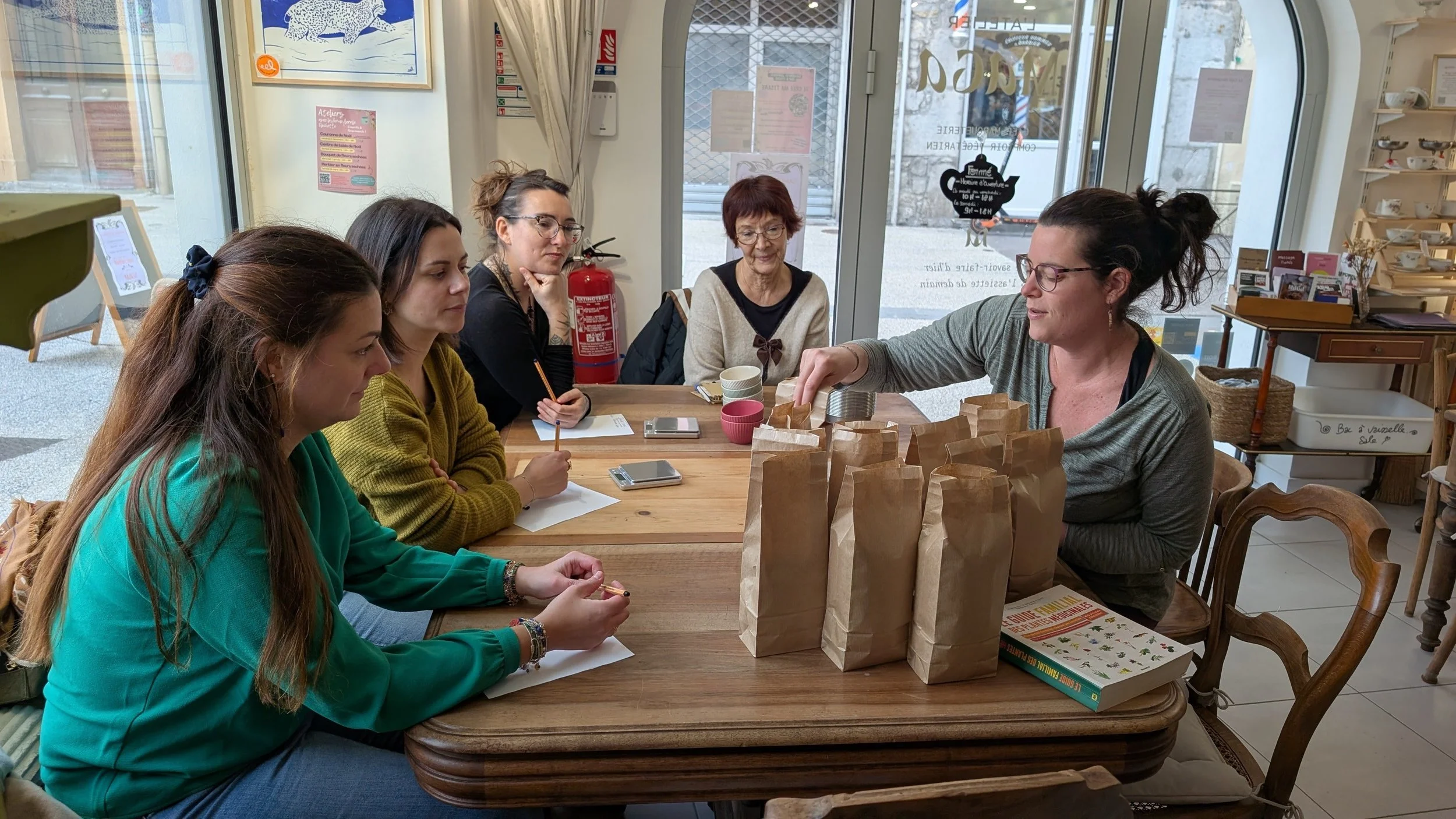 Un groupe de femmes assises autour d'une table en bois dans un café ou une boutique, regardant une femme qui prépare des sacs en papier devant elles.