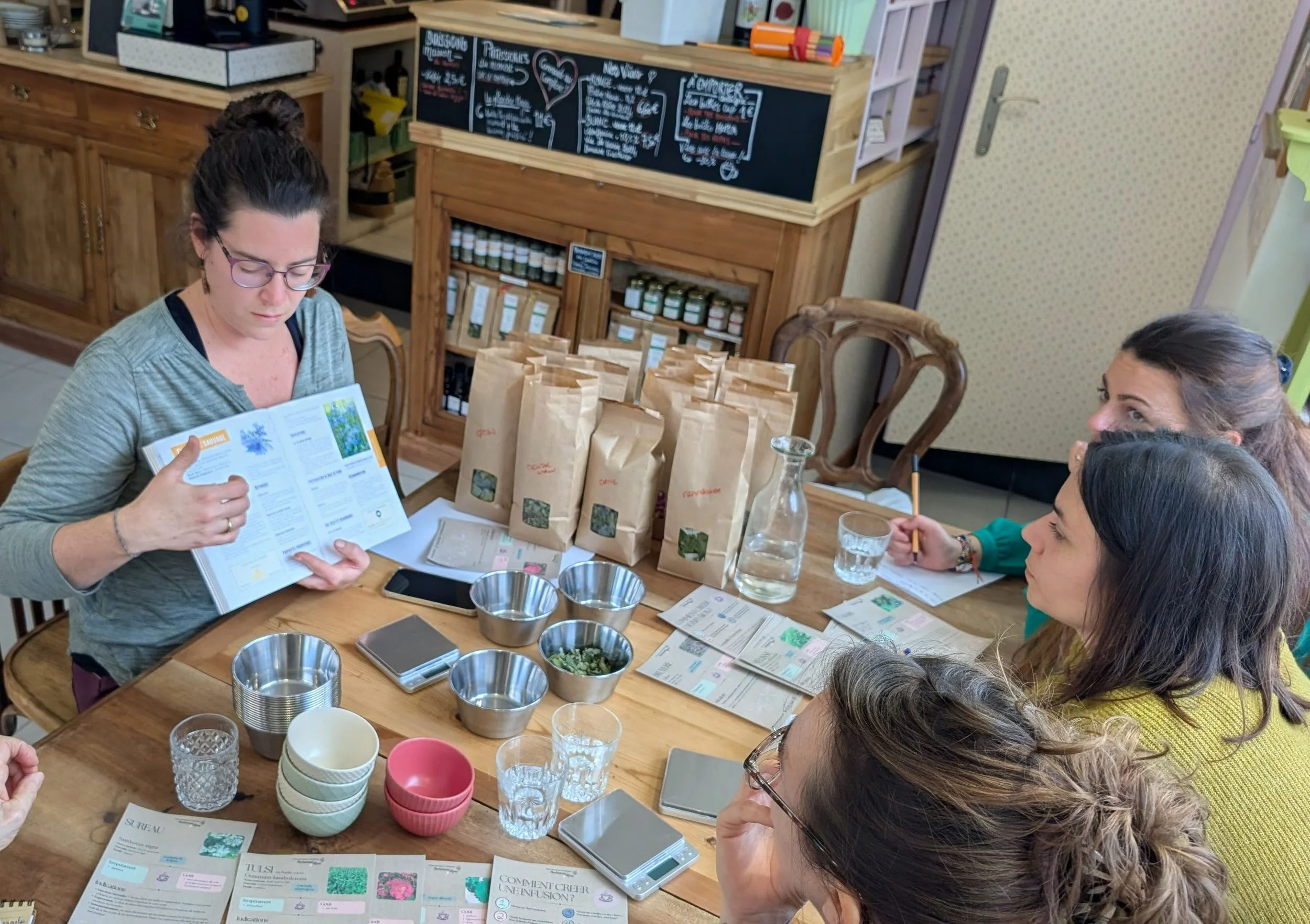 Groupe de femmes assises autour d'une table en bois, participant à un atelier de dégustation ou de présentation de produits bio ou naturels, avec sacs de produits, feuilles d'information, verres, bols et balances sur la table.