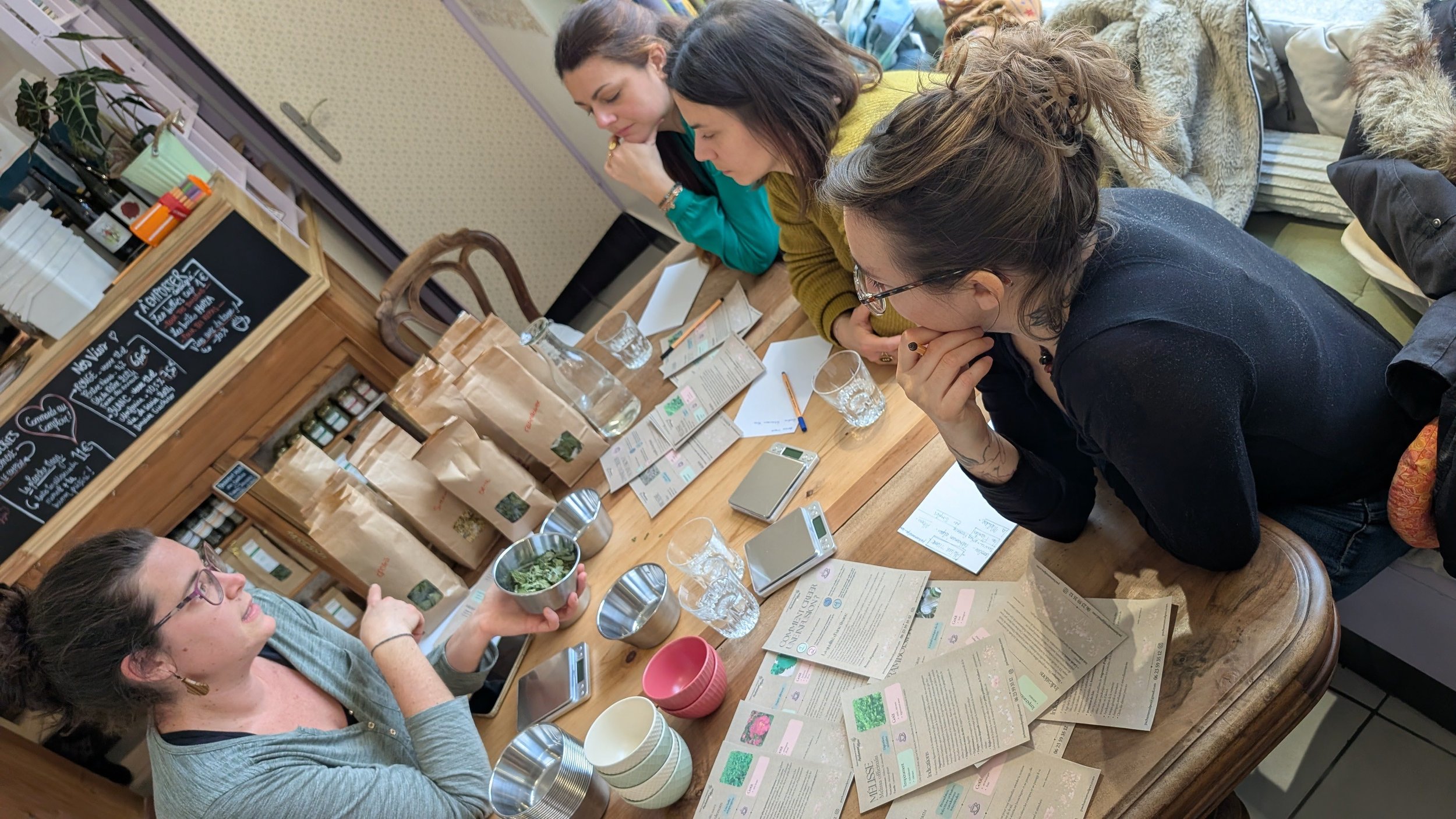 Groupe de quatre femmes discutant à une table avec des sacs en papier, des échantillons, des balances, de l'eau et des notes dans un café ou une boutique