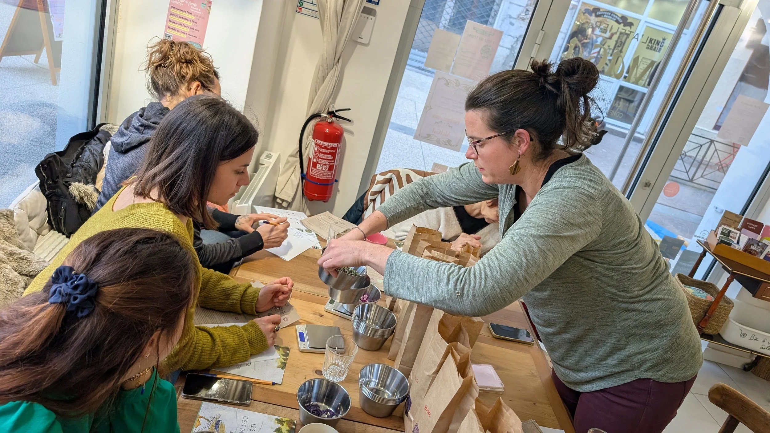 Une femme fait une dégustation ou une vente de produits à un groupe de personnes dans un café ou une boutique, avec des sacs en papier sur la table.