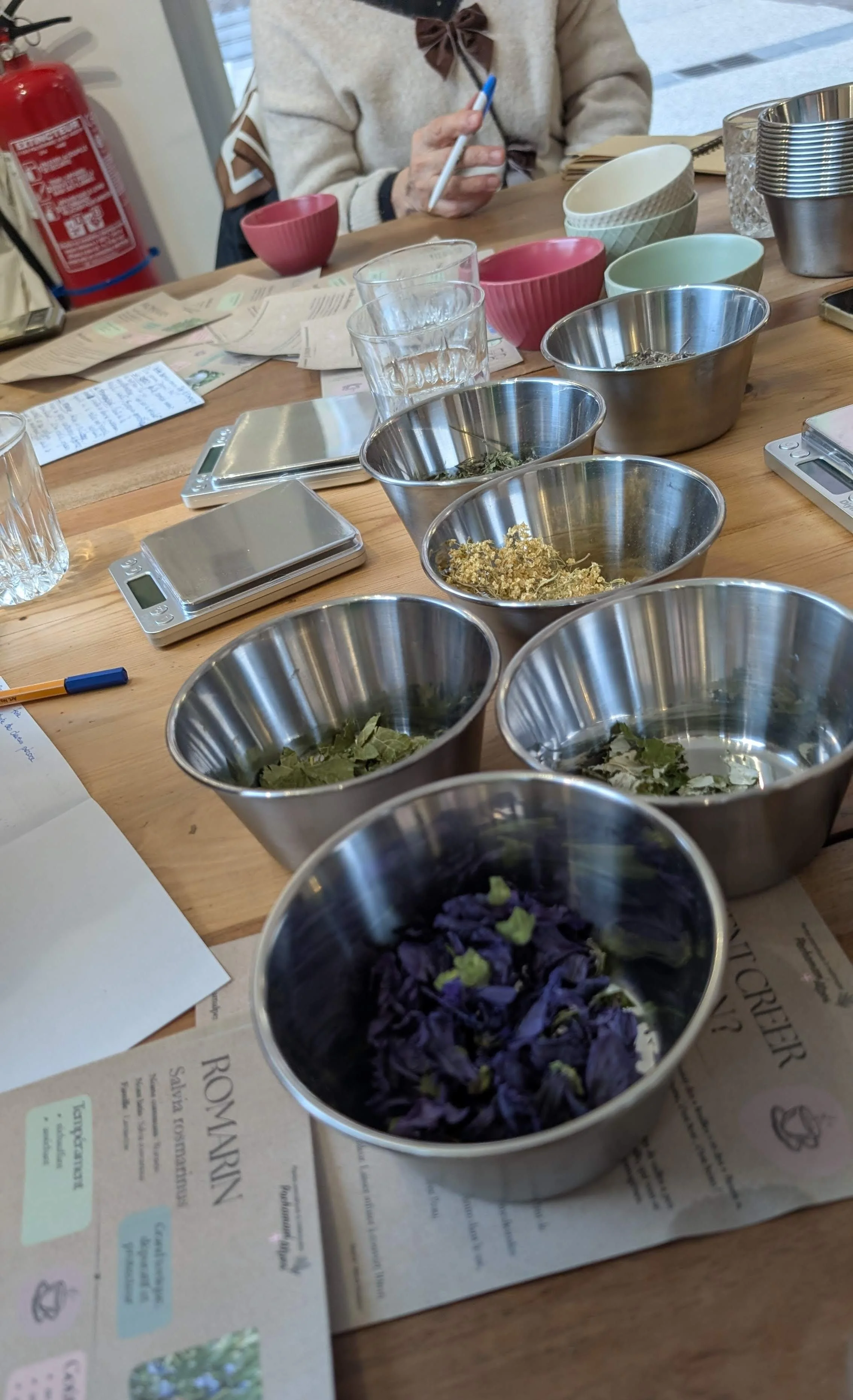 Tasses en acier inoxydable avec des fleurs séchées colorées, une bouteille d'eau, des papiers, une balance numérique, un stylo, une personne prenant des notes et quelques bols colorés et en métal, tout cela sur une table en bois.