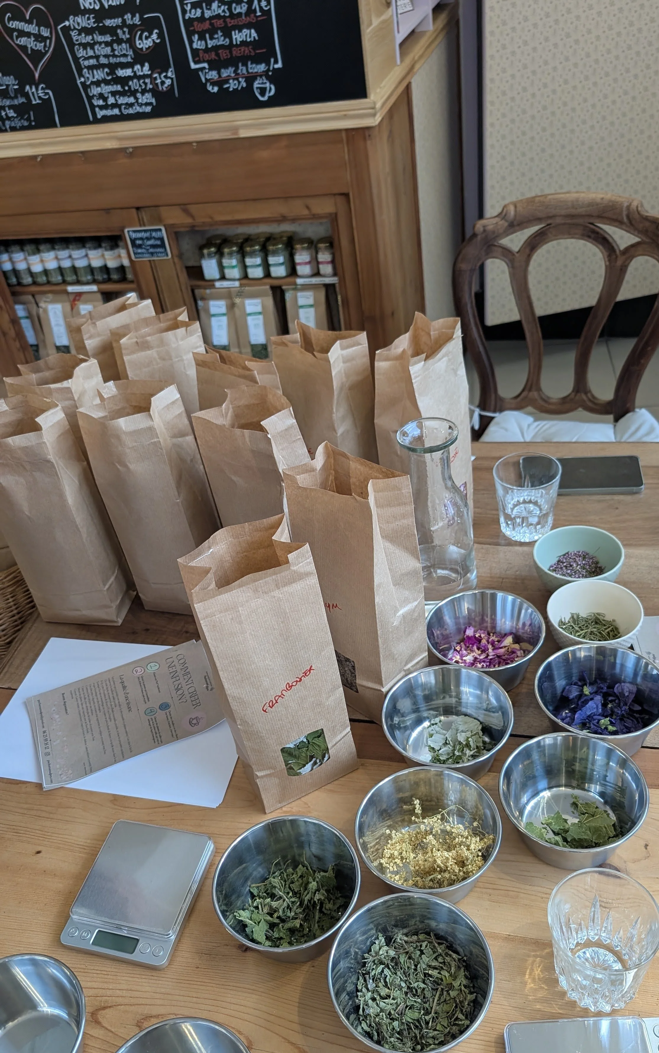 Table avec plusieurs sachets en papier kraft, bols avec différentes fleurs et herbes séchées, une balance numérique, un carnet, une bouteille en verre, un verre à eau, un smartphone, et un tableau noir avec un menu écrit en français.