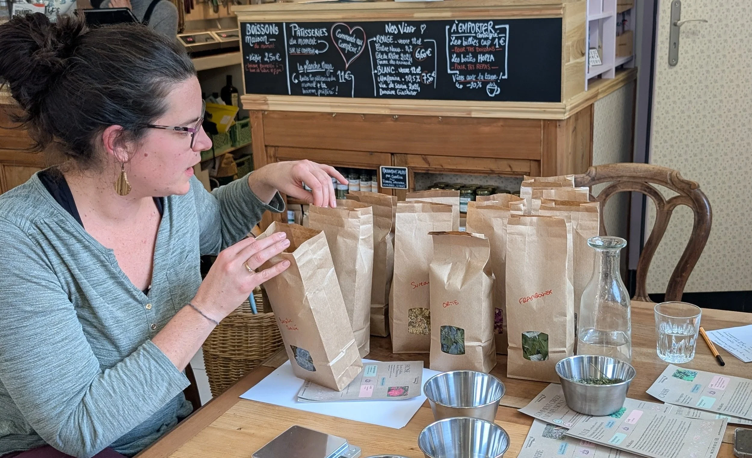 Femme examinant plusieurs sacs en papier contenant des herbes, sur une table en bois, dans une boutique ou un café, avec un tableau noir affichant un menu en français à l'arrière-plan.